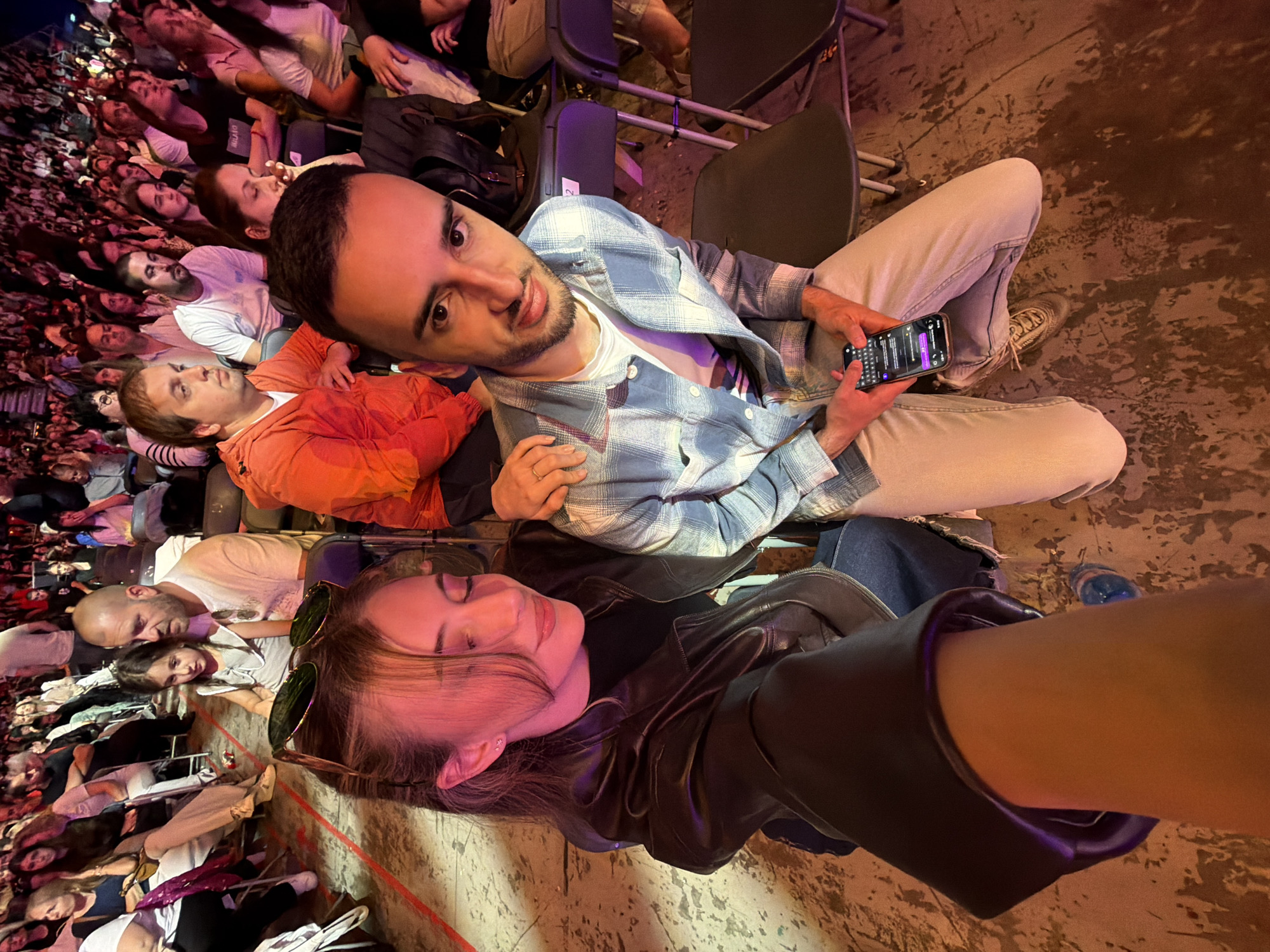 The image appears to be a selfie taken at an event, possibly a concert, in Sofia, Bulgaria. A woman wearing a black leather jacket lies in the foreground. Next to her sits a man holding a mobile phone. He wears a light blue plaid shirt. The screen of his phone shows a messaging app open. Behind the two individuals is a densely packed audience seated in rows of folding chairs. The flooring appears to be concrete.