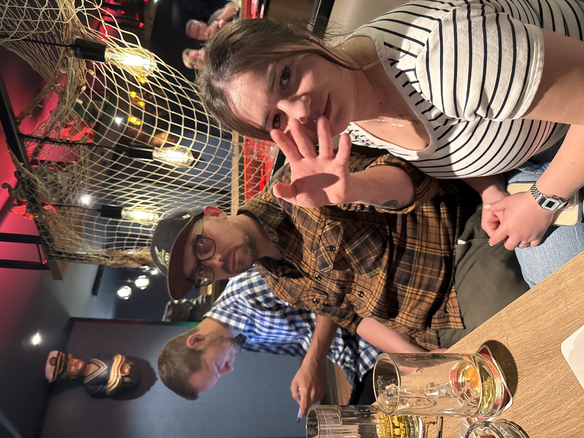 The image depicts an indoor scene at a bar or restaurant in Langeoog, Germany. In the foreground, a woman with brown hair and a striped shirt is looking towards the camera with her hand raised. To her left, a man wearing a plaid shirt and a baseball cap is also looking forward, with a beard and glasses. Behind him, another man in a blue checkered shirt is looking down at his phone. The environment is decorated with nautical-themed elements, including fishing nets and a bust of a sailor. There are beer mugs and a coaster visible on the wooden table in the foreground. The lighting is dim with warm tones.