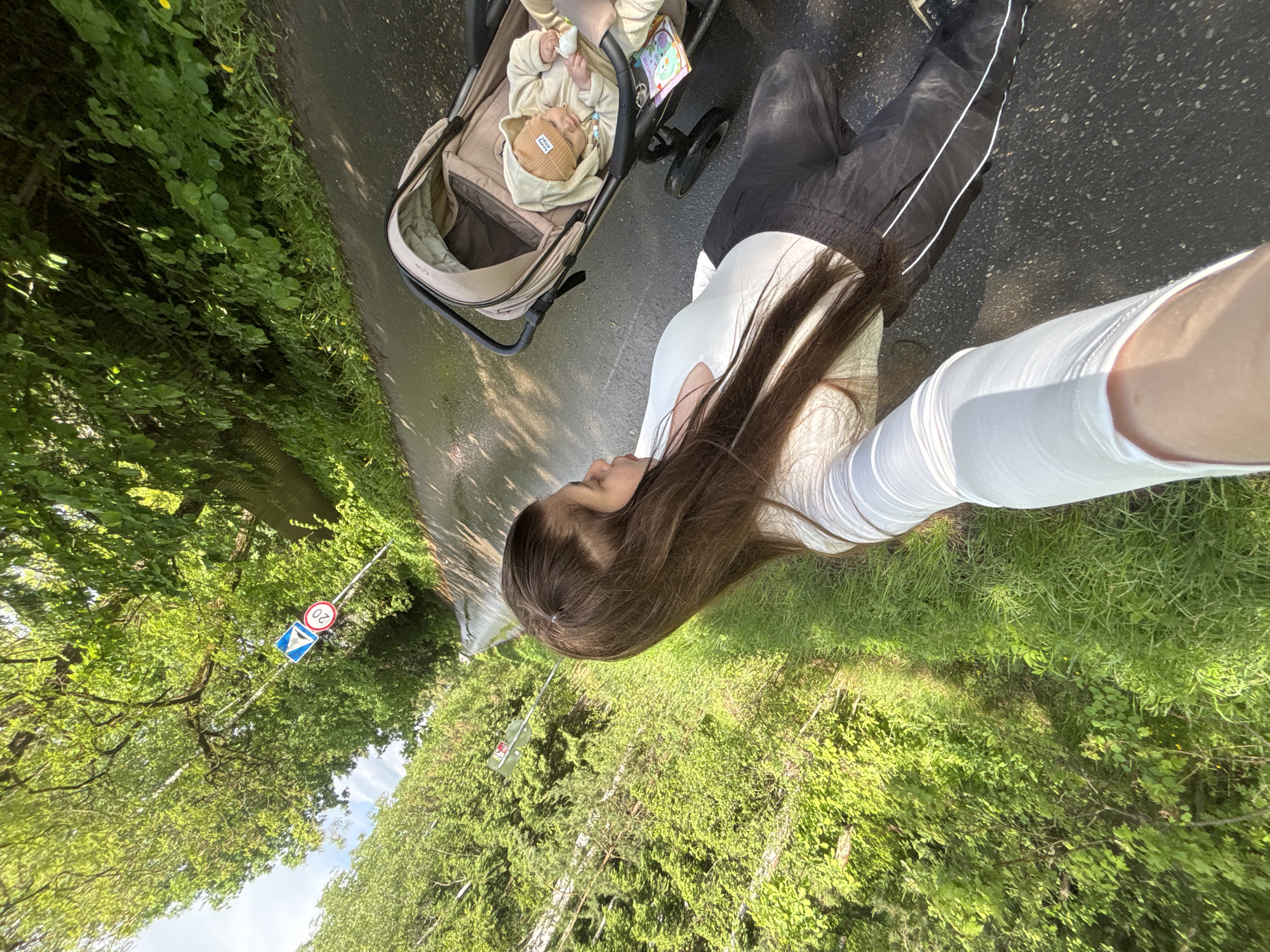 A woman is taking a selfie while pushing a baby stroller on a paved road in Shevlyagino, Russia. The stroller contains a baby wearing a beige outfit and hat. The woman is wearing a white top and black pants. A road sign indicating a speed limit of 20 km/h and a yield sign are visible on the left side of the road amidst trees. The environment is green and sunny.