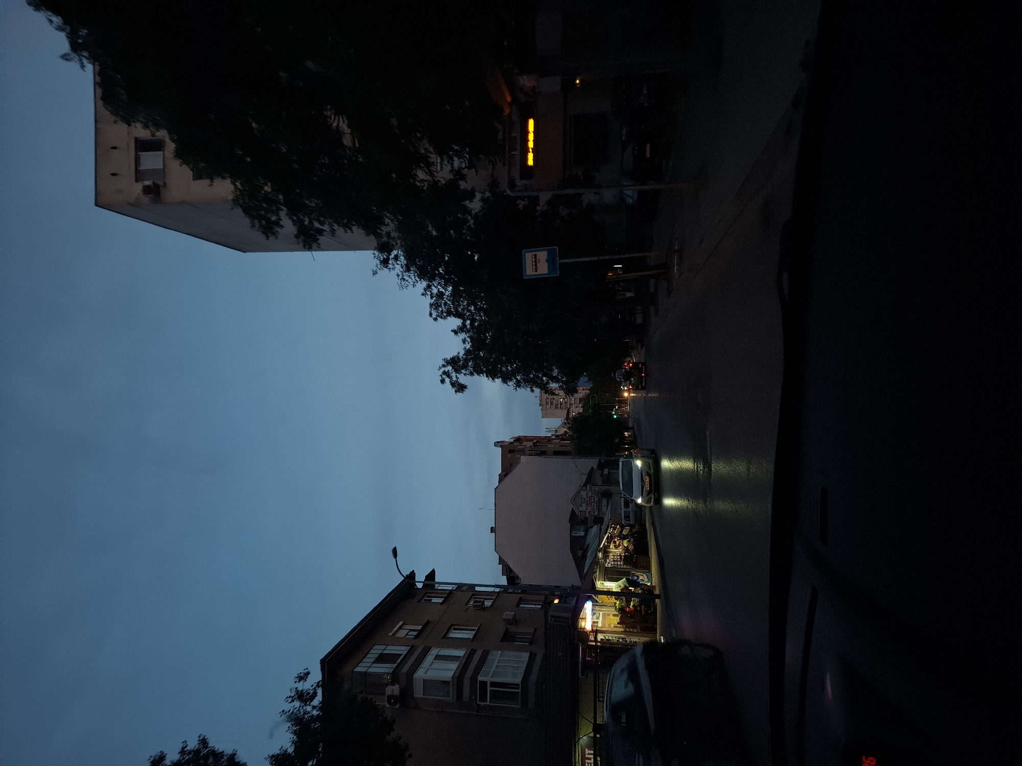 The image shows a street scene at dusk in a city. The sky is a deep blue, transitioning to dark as night falls. On the left side of the street, there is a multi-story building with several windows and balconies. Below the building, there are small shops with their lights on, displaying goods and one person working. A dark-colored car is parked in front of the shops.
On the right side of the street, there is another tall building, mostly obscured by dark trees. A bus stop sign is visible. Further down the street, traffic lights are on, and several cars are driving, their headlights illuminating the wet road. One car in the foreground has its headlights on, reflecting on the wet asphalt. The overall impression is a somewhat urban and commercial area as the day ends. The image appears to be taken from inside a vehicle.