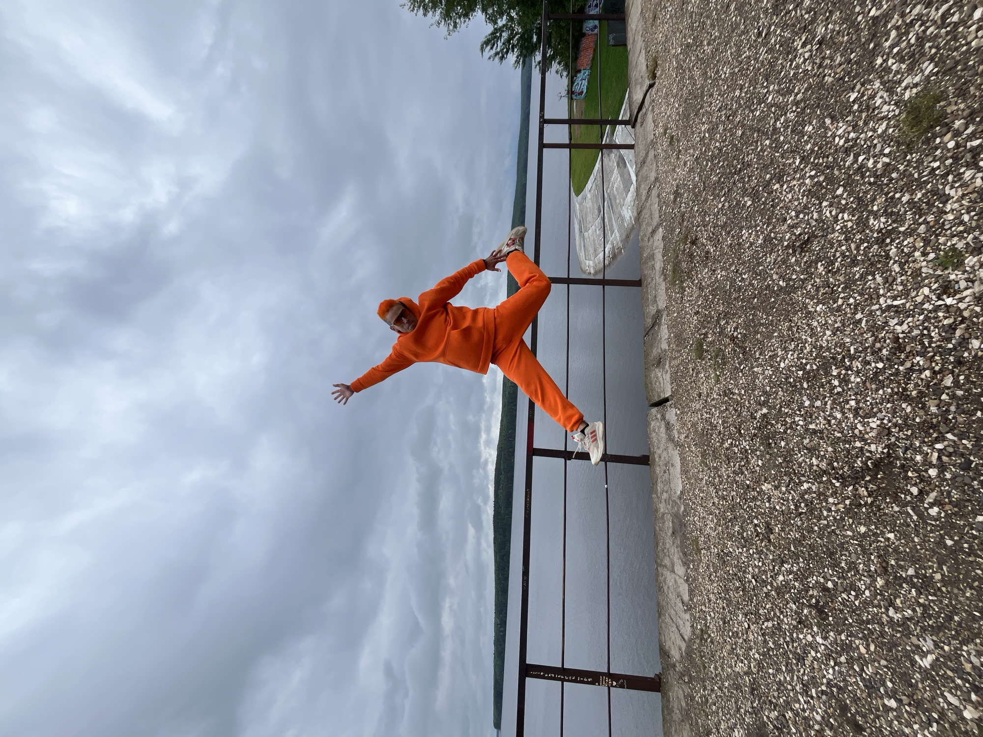 A person wearing an orange hooded tracksuit and white sneakers is performing a yoga pose on a metal railing. The railing is next to a concrete embankment along the Kama River in Chaikovsky, Russia. The sky is overcast. The embankment is made of concrete and stone aggregate. Trees and graffiti are visible behind the person and railing.