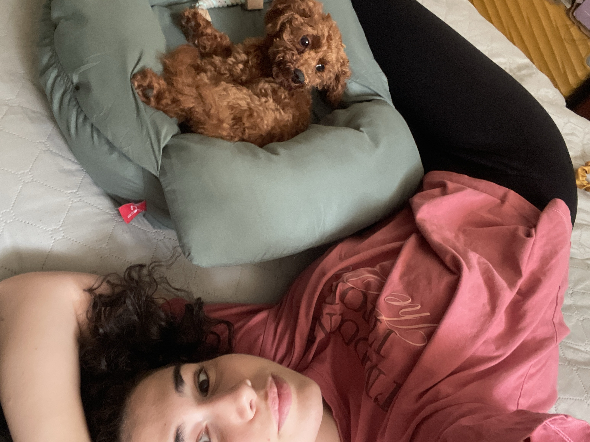 A young woman with curly dark hair lies on a bed, propped up against a green, round dog bed containing a reddish-brown poodle. She is wearing a rose-colored t-shirt and black leggings. The bed has a white quilted cover, with a yellow blanket visible in the upper right corner. The location is not identifiable as Sofia, Bulgaria from the provided image.