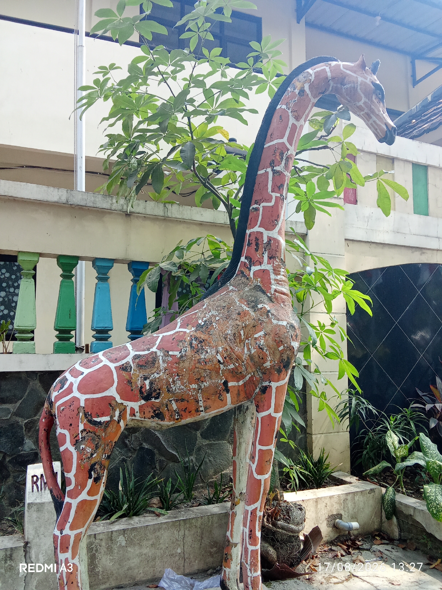 A statue of a giraffe, painted with orange and white spots, stands in the foreground. The statue is weathered and shows signs of age and damage. Behind the giraffe, a building with a pale yellow facade and a blue roof is visible. A balcony with colorful railings in green and blue overlooks a section of the building. Lush green foliage from a tree with large leaves partially obscures the building and the giraffe statue. To the right of the giraffe, a black, diamond-patterned wall and various plants create a textured background. The ground is a mix of concrete, dirt, and fallen leaves. A white pipe is visible near the base of the giraffe statue. The image includes a timestamp in the lower right corner, "17/08/2026 13:27," and the text "REDMI A3" in the lower left corner. The location is identified as Surakarta, Indonesia.