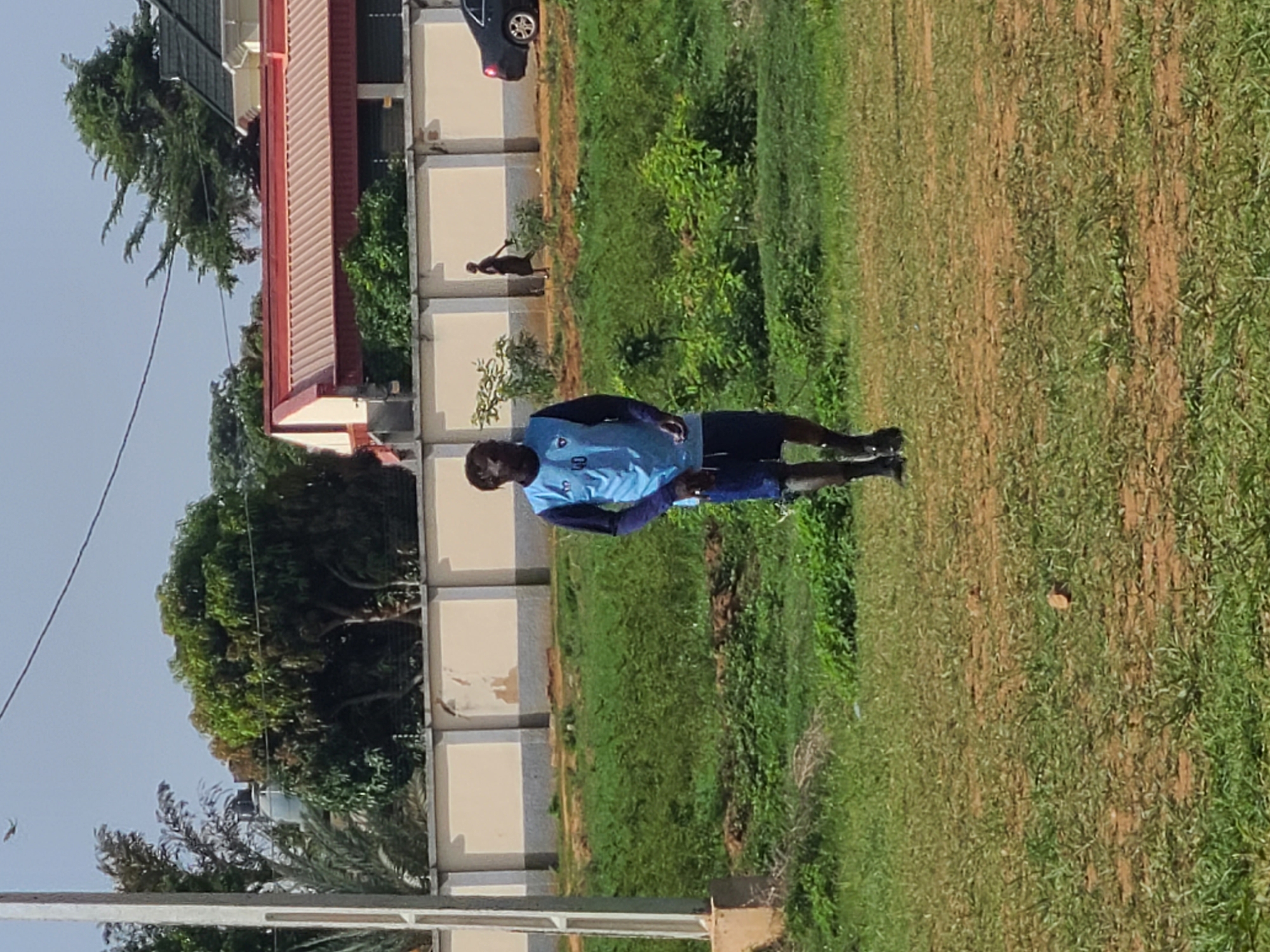 The image shows a man standing on a grassy field in what appears to be an urban environment. The man is wearing a light blue athletic shirt and dark shorts, and he is facing the right side of the frame. Behind him is a low concrete wall, and behind the wall are trees, a building with a red roof, and a car. The grass is patchy, with areas of bare dirt visible. The location appears to be in the city of Unknown, Unknown.