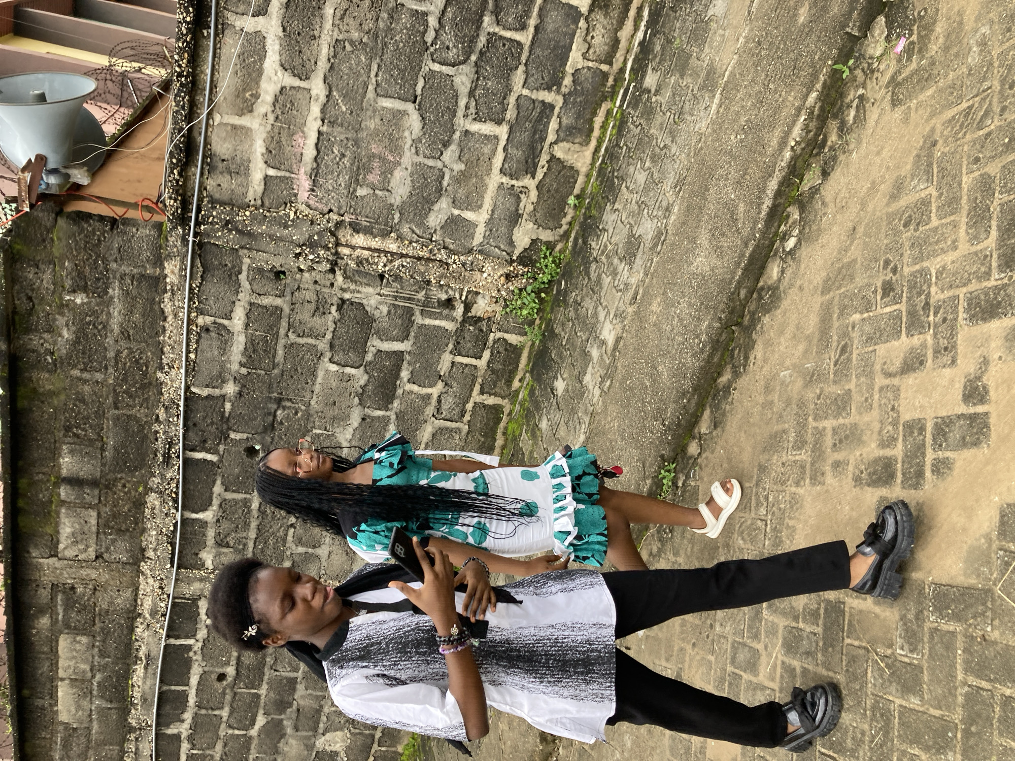Two young females are walking along a paved path next to a grey brick wall in Umuode, Nigeria. The female on the left, wearing a black and white patterned tunic, black pants, and black loafers with a bow, is holding a smartphone. The female on the right wears a green and white ruffled dress and white sandals. A large speaker is mounted on the wall behind them. The environment appears to be outdoors, with some vegetation growing along the base of the wall and on the paved ground.