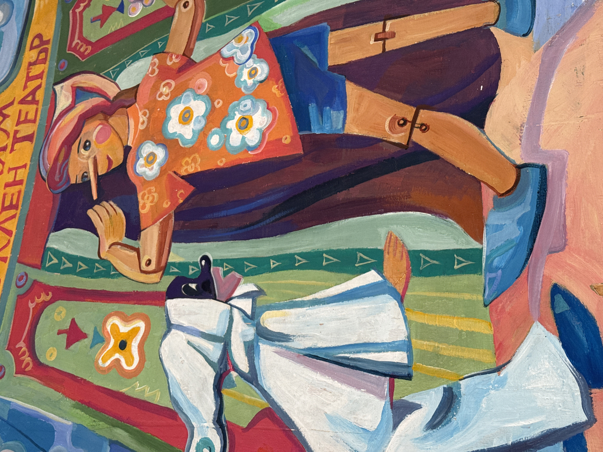 This vibrant painting depicts a scene from a puppet theater, characterized by its folk-art style and rich colors. The artwork features two distinct marionette-like figures against an elaborately patterned background.
The main subject is a wooden-jointed puppet, likely male, positioned horizontally across the upper portion of the frame. It is dressed in an orange shirt with white and light blue floral patterns, blue shorts, and light blue shoes. Its face features a prominent, long nose reminiscent of Pinocchio, along with rosy cheeks and wide eyes, all beneath a red and white striped hat. One of its hands is held near its face, possibly holding a small prop. The puppet's limbs clearly show the wooden construction and joint mechanisms, highlighted by brown straps and circular pivots.
Partially visible below and to the right is a second puppet figure, predominantly white with light blue and grey accents. This figure appears to be in motion, perhaps leaping or running, though only its lower body and a suggestion of its upper form are discernible.
The background is a colorful, patterned surface, resembling a theatrical stage floor or a decorative textile, like a carpet. It features bold geometric shapes and stylized floral motifs in shades of green, red, yellow, and blue, creating a dynamic and engaging setting. Along the left edge, a vertical yellow banner displays Cyrillic text: "ТРАЕН ТЕАТЪР". This translates to "TRAEN THEATER" or "LASTING THEATER," confirming the theme of a puppet performance and hinting at the location context of Sofia, Bulgaria. The painting style is expressive, utilizing bright hues and distinct brushstrokes to create a whimsical and narrative atmosphere.
