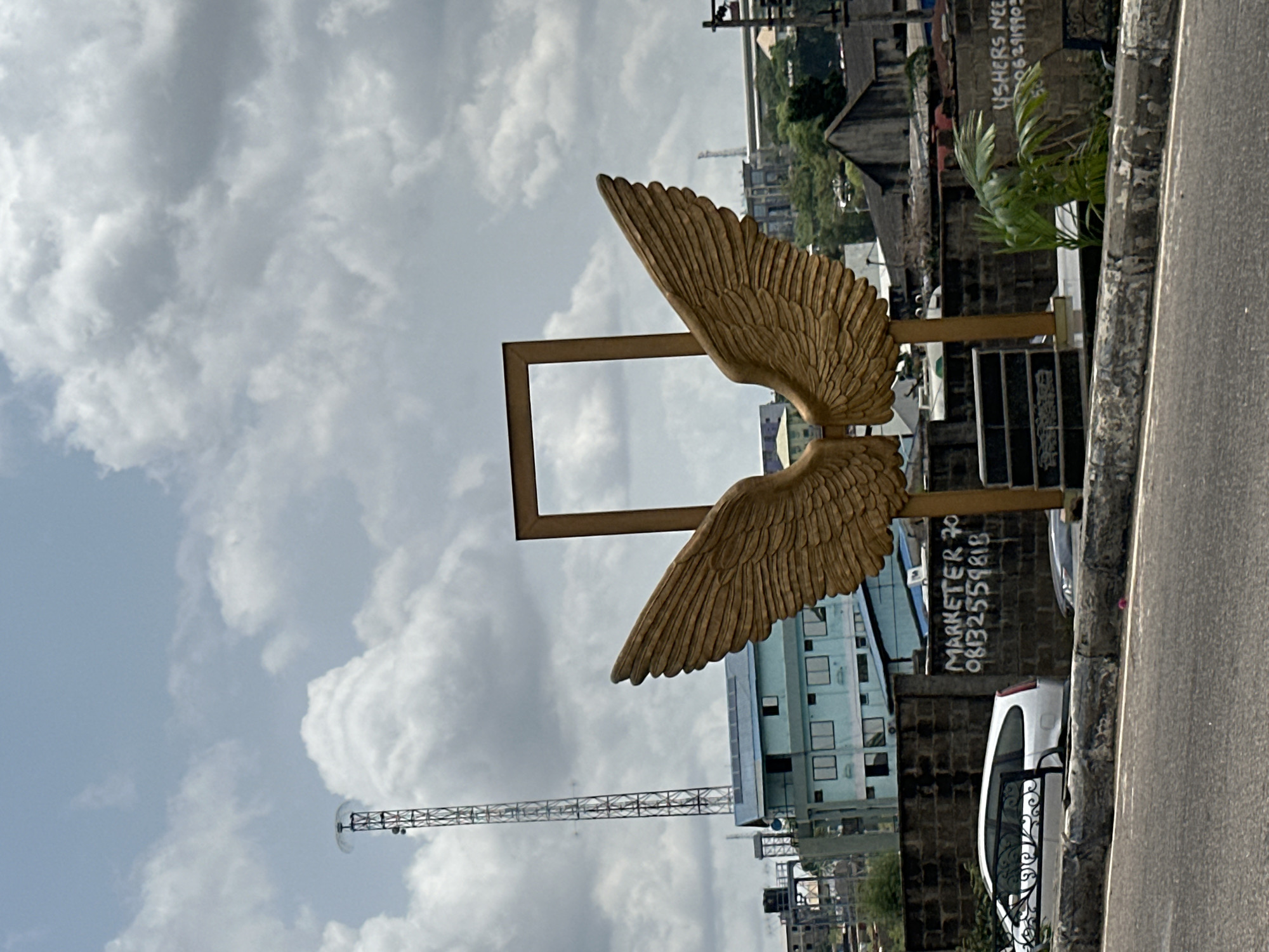 A photograph captures a sculpture in Ikeja, Nigeria, featuring a pair of golden wings within a rectangular frame. The sculpture is set against a backdrop of buildings and a cloudy sky. A communications tower rises in the distance. A low wall with graffiti including "MARKETER 70 08132559818" borders a street where a white car is parked. Lush greenery grows nearby.