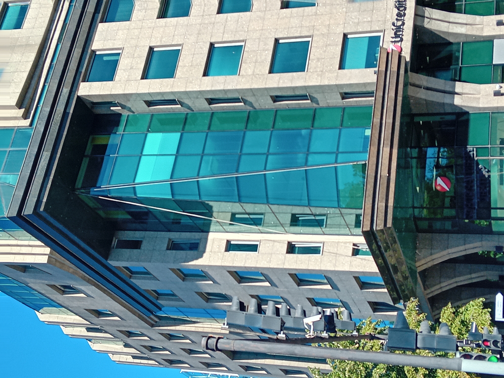 The image depicts the UniCredit Bank building in Varna, Bulgaria, on a clear day. The building is a multi-story structure with a modern architectural design, characterized by a combination of stone and glass facades. The stone facade has a textured, light-colored finish with visible rectangular blocks. The glass facade is predominantly turquoise blue, with some sections of lighter green.
On the left side of the image, a traffic light pole with multiple signals is visible, with one green light illuminated. A traffic sign with white arrows on a dark background is also present at the bottom. Trees with green foliage are seen partially obscuring the lower part of the building and the traffic signal pole. The building's entrance area features a prominent overhang with a glossy dark surface and decorative stone pillars. The UniCredit logo, a red circle with a white stylized curve, is visible on the building's facade and reflected in the glass. The sky is a clear, bright blue, indicating good weather.