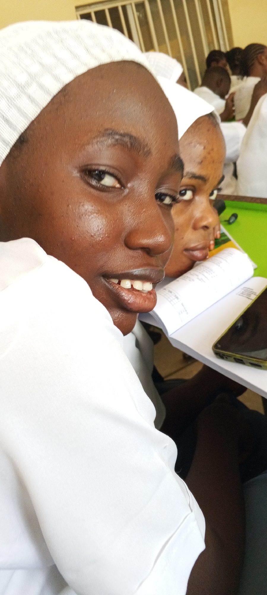 A close-up, eye-level shot shows two young women seated at a green table in Keffi, Nigeria. The foreground woman, of African descent, is smiling slightly. She wears a white, collared shirt and a white head covering with a subtle grid pattern. The woman behind her, also of African descent, is looking directly at the camera with a neutral expression. A book is open between them, and a smartphone rests on the table. In the blurred background, more individuals wearing white clothing are visible. A window with vertical blinds is slightly visible in the upper left corner.