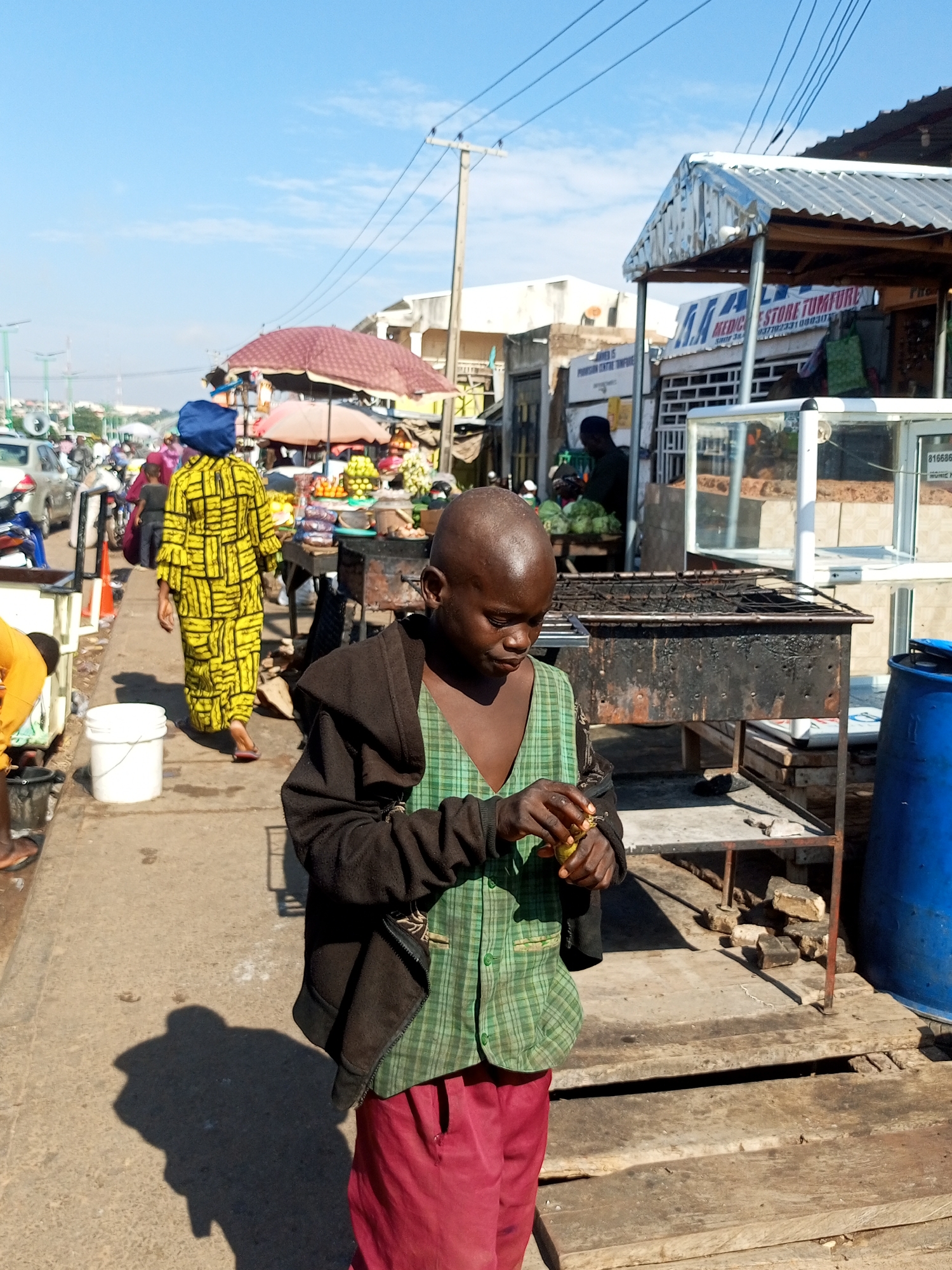 In the city of Tempure, Nigeria, on a sunlit day, a young boy with a shaved head stands near a food stall. He wears a green plaid vest over a dark jacket and red pants. He holds a piece of food in his hands, seemingly peeling or preparing it. To his right, a large blue barrel is visible. Behind him, a charcoal grill and a display case with goods are set up at a market stall. Further in the background, other market stalls, people, and vehicles are present. A woman in a vibrant yellow and black patterned dress walks away from the camera towards the stalls, her blue headscarf noticeable. The scene depicts a bustling outdoor market environment.