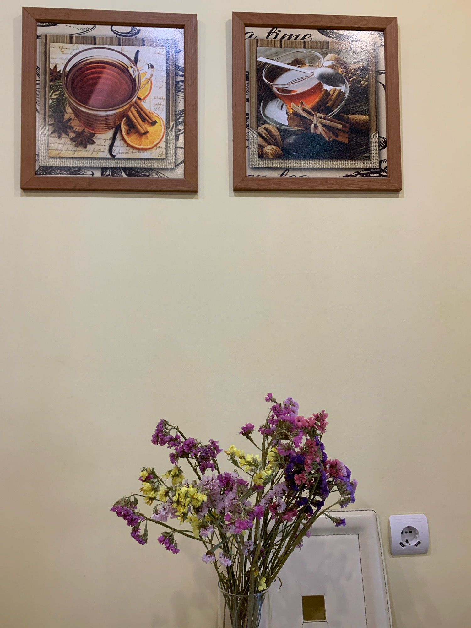 The image shows a wall with two framed pictures of tea and a vase of dried flowers. The pictures, hung side by side, feature close-ups of tea in glasses with cinnamon sticks and dried oranges. Below the pictures, a clear glass vase contains a bouquet of dried flowers in shades of purple and yellow. To the right of the vase, a white electrical outlet with two USB ports is visible on the wall. The overall setting appears to be indoors, possibly a cafe or home, given the decorative elements. The lighting suggests an indoor environment, with a neutral-colored wall as the backdrop. The presence of an electrical outlet with USB ports is a modern detail. The image does not contain any people. It is not possible to determine the location within the city of Minsk, Belarus from the provided image.