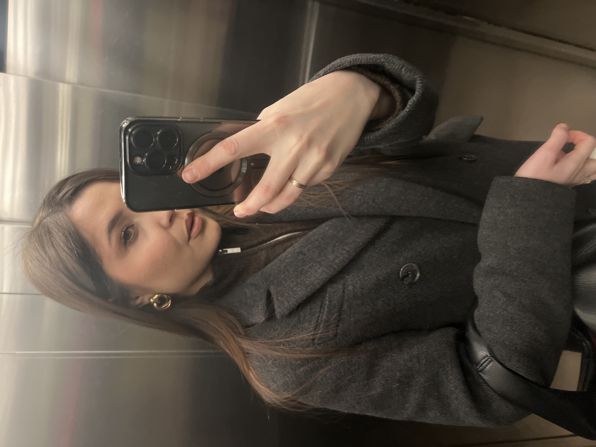 A young woman is captured in a mirror selfie inside what appears to be an elevator with metallic, reflective walls in Khimki, Russia. The image is oriented sideways, showing the woman's head tilted towards the left, looking directly into the camera of her smartphone.
She has long, brown hair and is wearing a dark grey or charcoal-colored coat over a darker top, possibly a sweater, which has a zipper visible underneath the coat lapel. A gold-colored hoop earring is visible on her right ear, and a thin gold-colored ring adorns her right ring finger. She holds a black smartphone, likely an iPhone due to its distinct triple camera array, encased in a clear cover, with a visible circular MagSafe accessory on its back. Her nails are short and unpainted.
Her expression is neutral, with her lips slightly parted. Her left hand is partially visible, holding a small white object, possibly a tissue or paper. A black leather-like strap, potentially from a bag, is seen near her left arm and shoulder. The background is a plain, brushed metallic surface characteristic of elevator interiors. No specific time of day or weather conditions can be discerned from the indoor setting, and there is no visible text in the image.