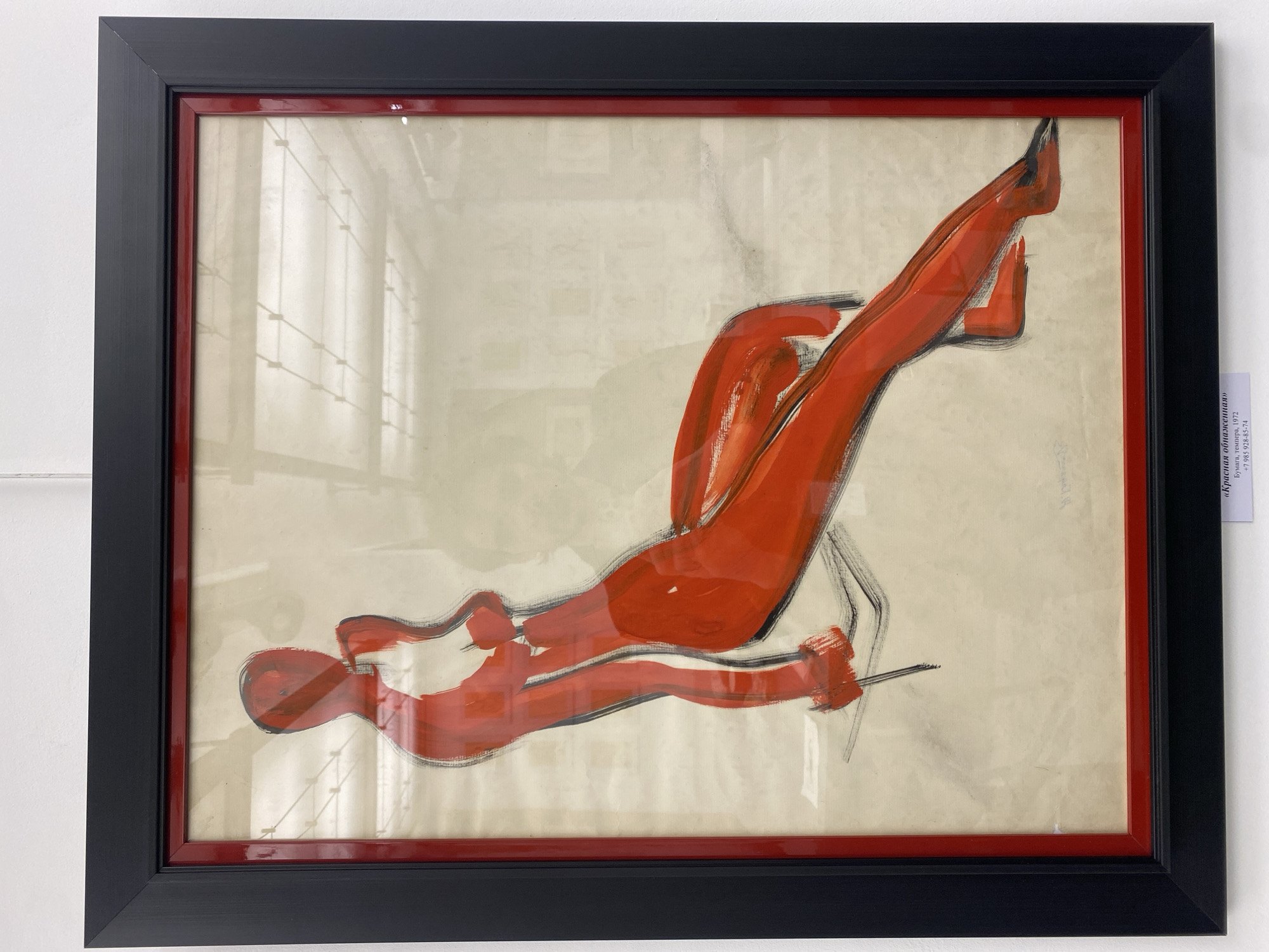 A framed artwork is mounted on a white wall. The artwork depicts an abstract human figure rendered in red brushstrokes with black outlining details. The figure appears to be reclining in an ambiguous posture. The frame consists of an outer black layer and an inner red layer. To the right of the framed artwork, a narrow vertical sign with Russian text is visible. The sign contains information regarding the artwork, specifying "бумага, темпера, 1972" which indicates paper, tempera, and the year 1972, along with contact phone number "+7 985 928-85-74." The setting is assumed to be Moscow, Russia, based on the Russian text and the presence of Russian phone number.