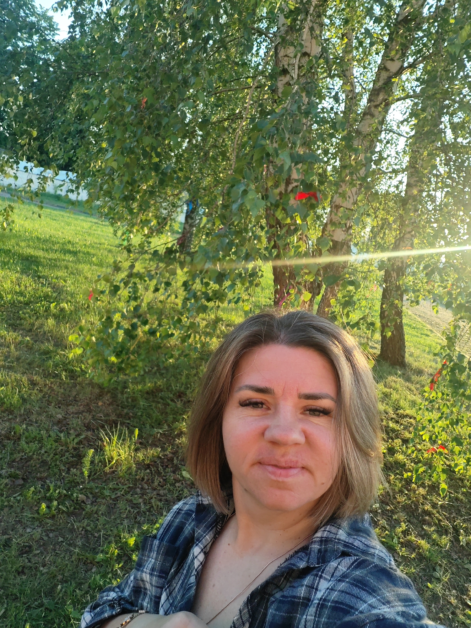 This media file captures a woman taking a selfie outdoors in Bronnitsy, Russia, during what appears to be the golden hour of either late afternoon or early morning. Warm, low sunlight filters through the trees, creating a prominent lens flare on the right side of the image.
The woman, with medium-length light brown hair and a neutral to slightly smiling expression, is wearing a blue and white plaid shirt. Her face is directly illuminated by the sun. The background reveals a natural setting dominated by several birch trees, identifiable by their distinctive white bark and abundant green leaves. Notably, multiple red ribbons are tied to the branches of these trees. The ground is covered in lush green grass, suggesting a park, garden, or rural area. The weather appears clear and sunny, contributing to the bright and natural feel of the scene. No text is visible in the image.