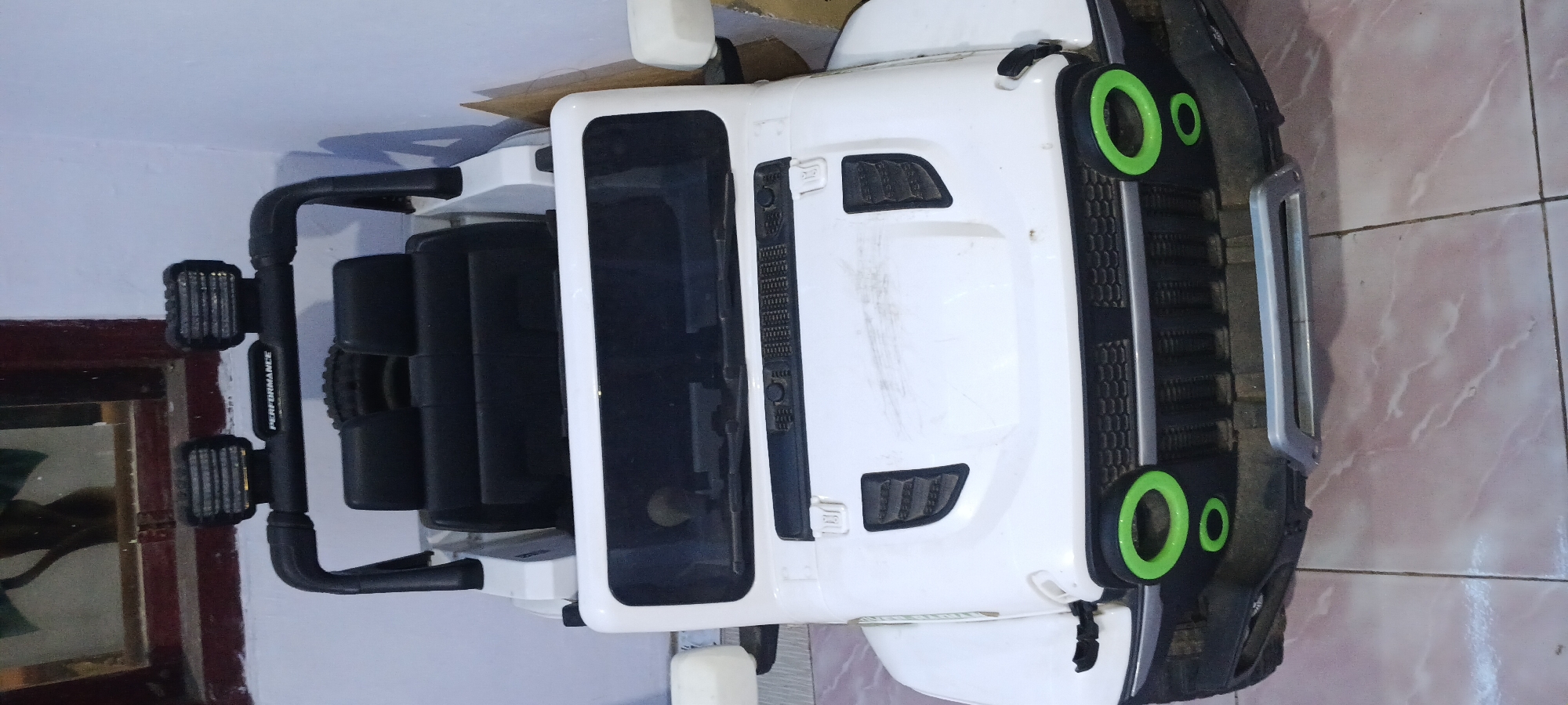 A white, toy Jeep Wrangler is parked on a tiled floor indoors, possibly in a residential setting in Rejing, Indonesia. The toy Jeep is front-facing and occupies the majority of the frame. It features two black seats, a black roll cage with two square headlights, and a front grille with green accents around the headlights. The exterior of the Jeep shows some scuff marks and dust. Behind the Jeep, a wall is visible, and to the left, a portion of a window with dark framing and reflections of greenery can be seen. The lighting appears to be artificial, casting soft shadows. There are no people or animals present in the image.