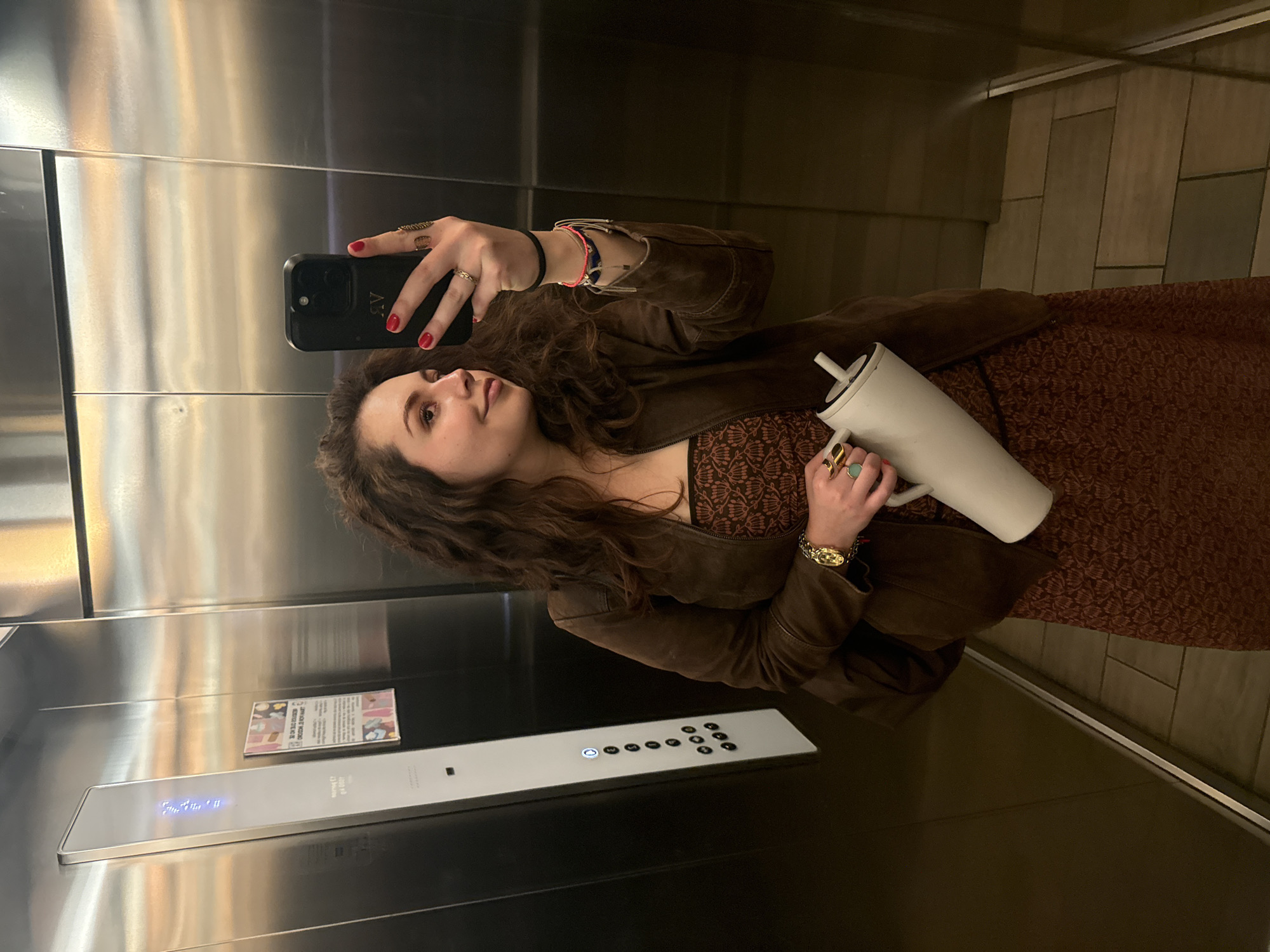 A woman is taking a selfie in an elevator. She is holding a black cell phone in her right hand, which is angled upward, with her left arm wrapped around a white object. The woman has long, curly brown hair, and she is wearing a brown jacket, and a brown dress with a brown pattern. She also has red nail polish. The elevator is modern, with stainless steel walls and a panel of buttons. There is a small sign on the elevator wall. It is unknown where in Mexico City, Mexico, this image was taken.