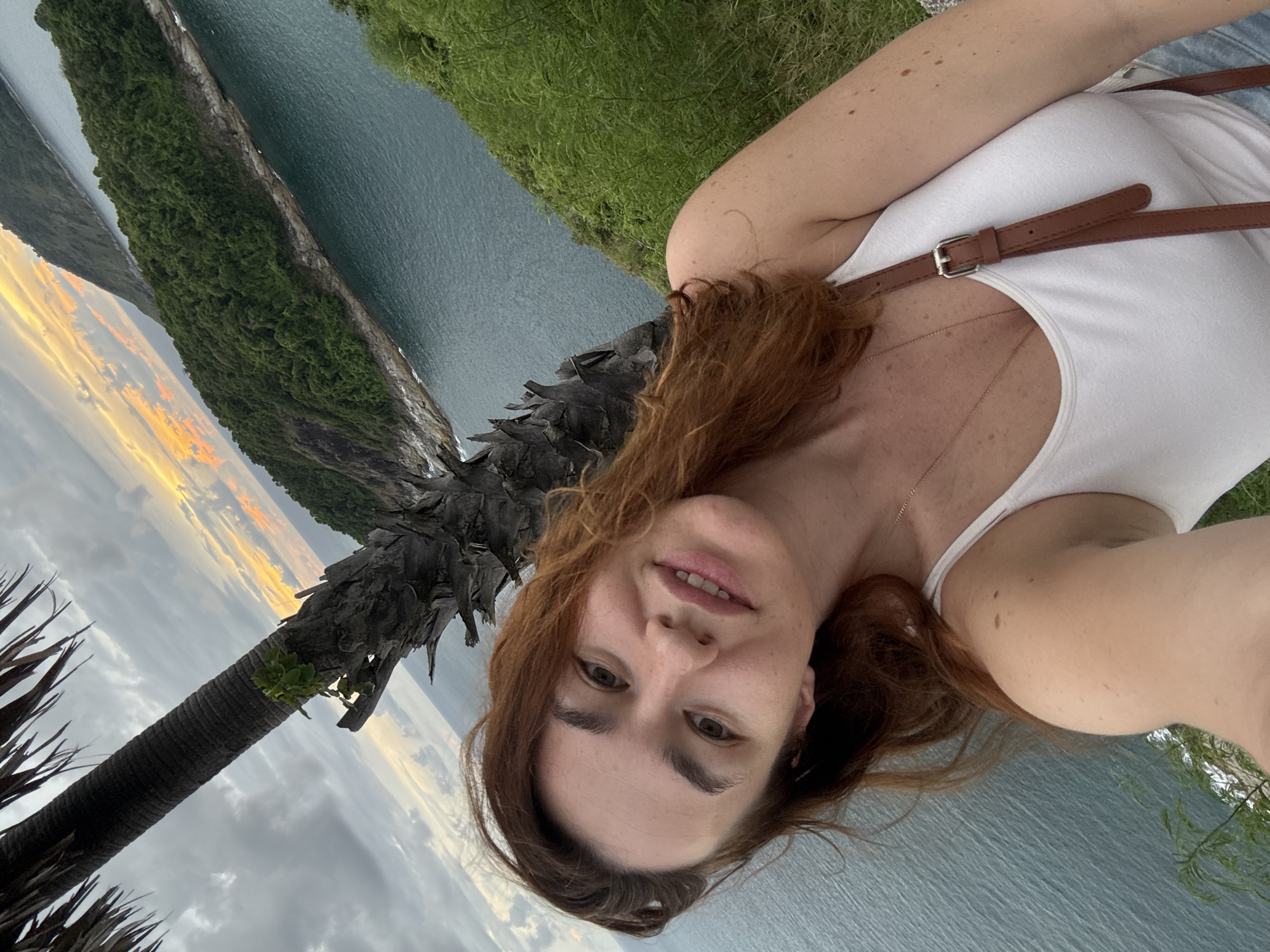 A woman with auburn hair is taking a selfie from an elevated vantage point overlooking a body of water and a tree. She is wearing a white tank top and a brown crossbody bag. The background shows a coastline with lush green foliage and a cloudy sky with hints of sunset colors. The image appears to be taken in Rawai, Thailand, indicated by the tropical vegetation and coastal scenery.