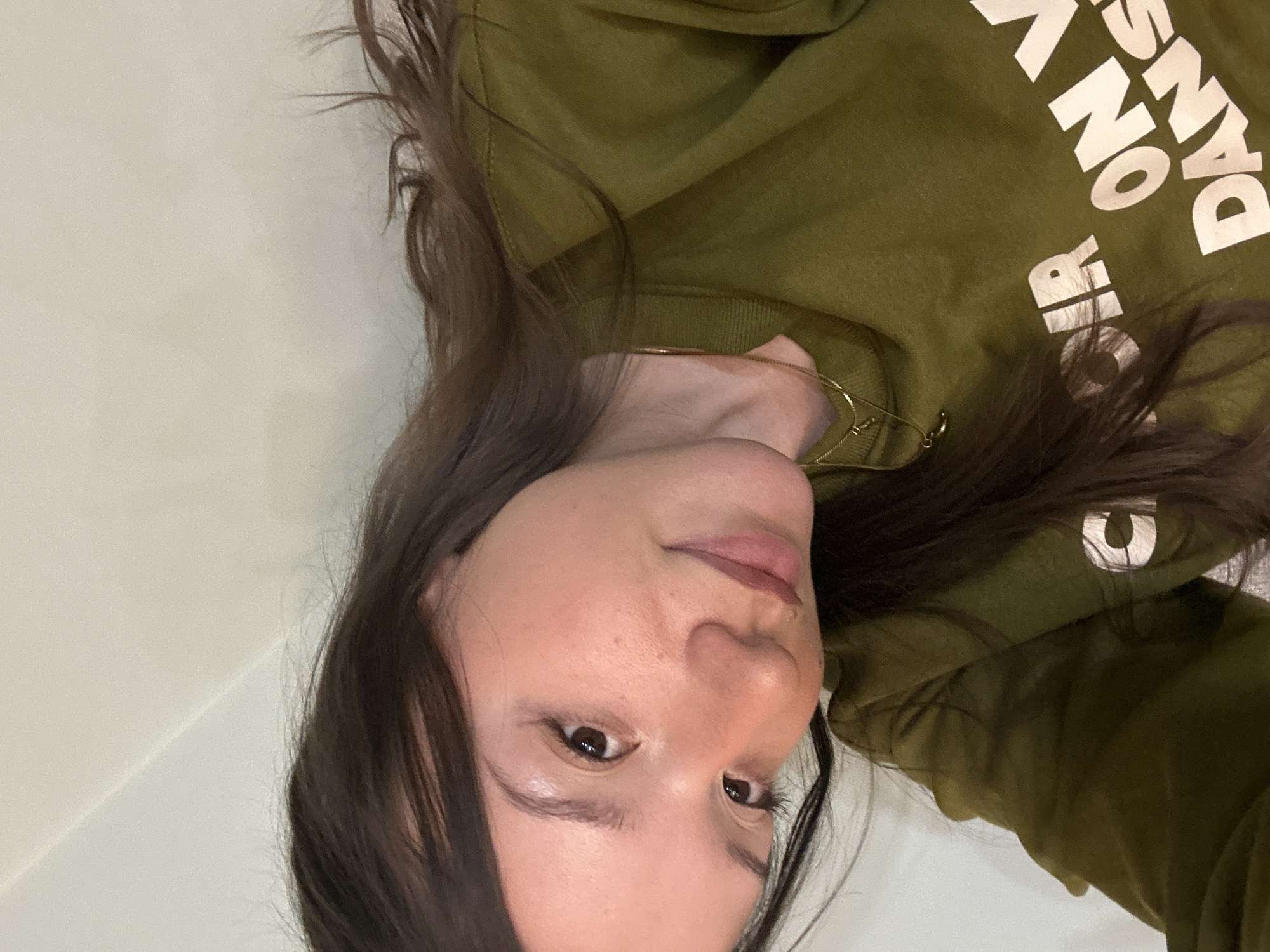 A woman with long, dark brown hair is facing the camera, wearing a green sweatshirt with white text that includes the words "ON DANSE." She is also wearing a gold necklace. The background appears to be a plain, white wall. There is no information to indicate a location in Sofia, Bulgaria, or any specific city.