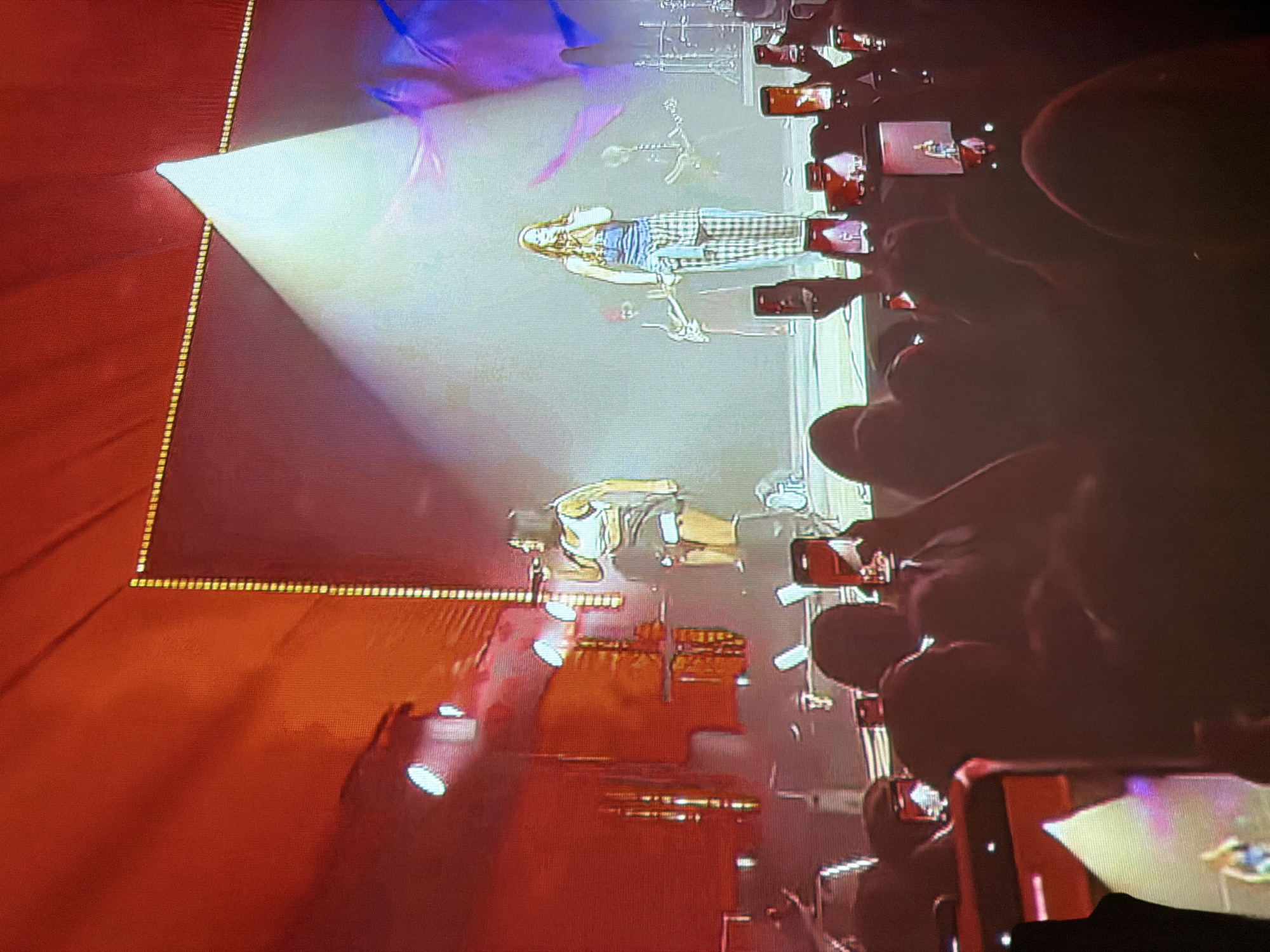 The image depicts a concert in an indoor venue. In the foreground, a crowd of concertgoers is visible, many holding up smartphones to record or photograph the performance. The stage area is illuminated by bright spotlights and a large screen. Two female performers are the central focus on stage. The performer on the left, with dark hair, is wearing a white crop top and a dark skirt. The performer on the right, with long, light-colored hair, is wearing a blue top and plaid pants. Several musicians are also visible on stage, playing instruments. The overall atmosphere appears energetic and engaged.