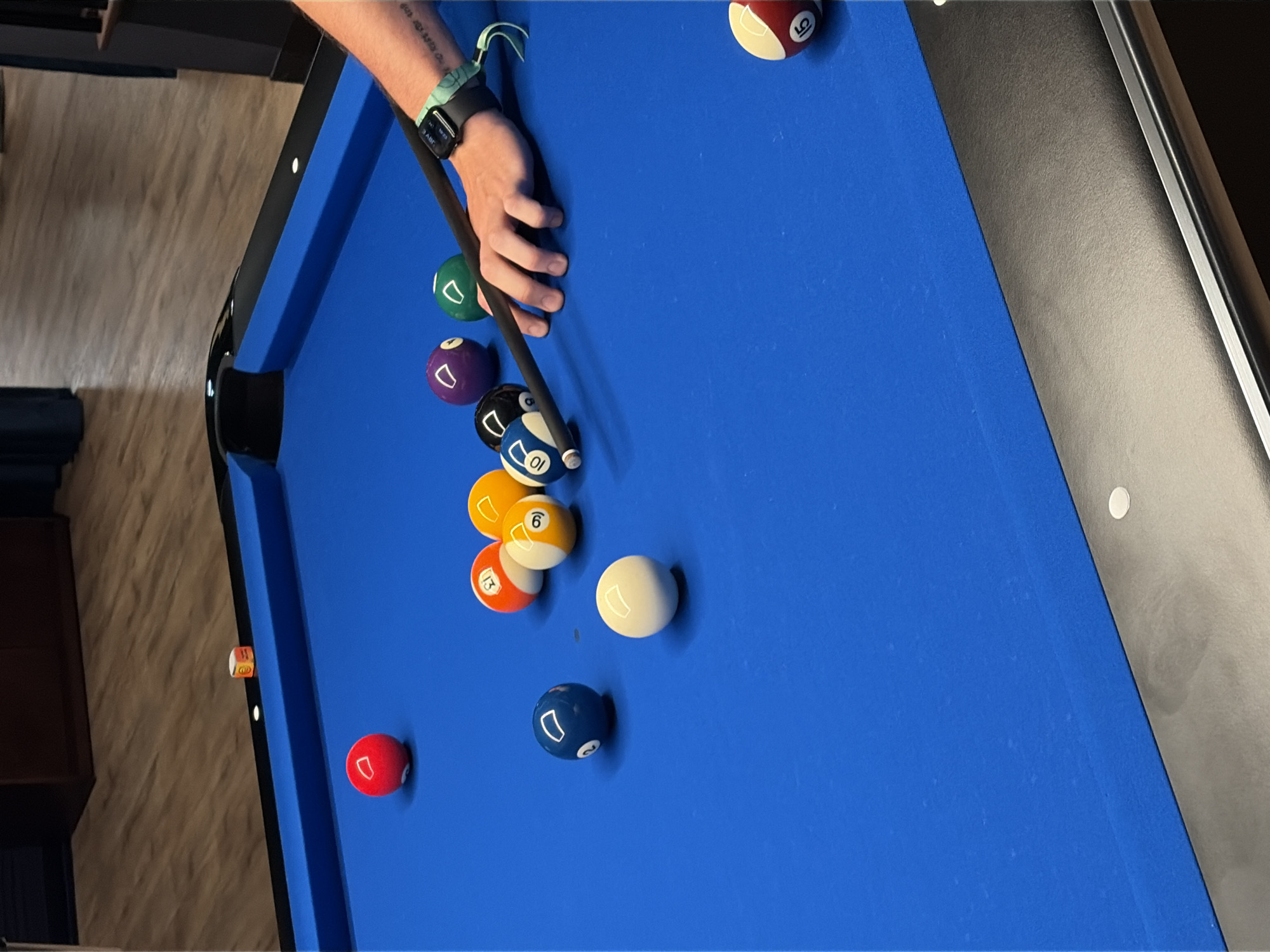 This is a close-up, eye-level, medium shot of a person playing billiards on a blue pool table. The pool table is well-maintained, with a vibrant blue felt and black rails. Several billiard balls are scattered across the table, including a solid red ball, a solid white ball, a blue ball with the number 2, a yellow ball with the number 11, an orange ball with the number 13, a yellow and white striped ball with the number 6, a black ball with the number 9, a purple ball with the number 3, a green ball with the number 12, a blue and white striped ball with the number 10, and a red and white striped ball with the number 15. A cue stick is being held by a person's hand, positioned to strike one of the balls. The person's arm and wrist are visible, and they are wearing a smartwatch. In the background, the wooden floor and a dark blue curtain are visible. There is also a small cube with orange writing on it placed on the rail of the pool table. The overall impression is one of a casual game of billiards being played in an indoor setting.
The image does not contain any elements that would allow for determination of its location in Moscow, Russia.