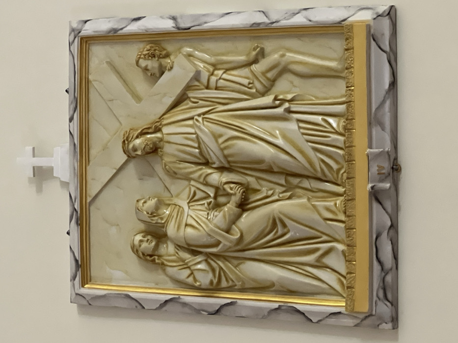 This image presents a rectangular relief sculpture mounted on a plain, light-colored wall in Nha Trang, Vietnam. The sculpture, rendered in a uniform light beige material, depicts the Fourth Station of the Cross, indicated by the Roman numeral "IV" centered on its lower border. The scene features four figures. The central figure, portraying Jesus Christ with a beard and long hair, carries a large cross over his right shoulder, looking towards the left. To his immediate left, a female figure (likely Mary) holds his left hand with both of her hands, gazing up at him. A second female figure stands further left, also looking towards the central figure. To the right of Jesus, a male figure, partially obscured by the cross, appears to assist with its burden, looking towards Jesus. The relief is enclosed within a multi-layered frame consisting of a thin golden inner border, a golden base plate, and an outer frame with a grey and white marbled pattern. Directly above the top center of the framed sculpture, a small, white, three-dimensional Latin cross rests on a small rectangular base, also affixed to the wall.