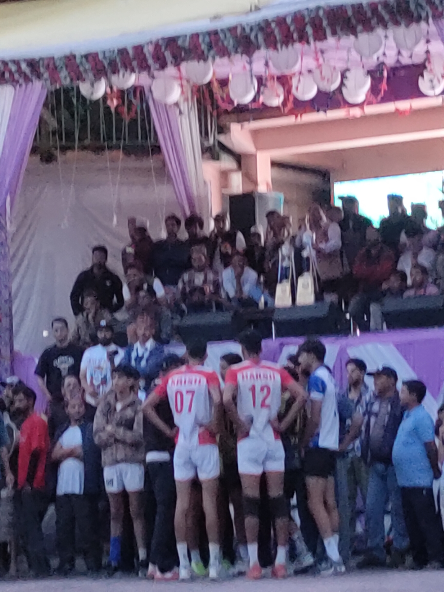 In Burwa, India, a group of individuals are gathered, possibly for an event or competition. The foreground features two young men wearing athletic attire. The person on the left is wearing a white and red jersey with the number "07" and the name "HARSH" on the back, accompanied by white shorts and knee pads. To their right, another young man wears a blue and white jersey with the number "12" and the name "HARSH" also visible. He is wearing black shorts.
Behind them, a diverse crowd of onlookers is present, some seated and others standing. They are dressed in a variety of casual clothing. The backdrop consists of decorative elements, including purple and white drapes, and what appear to be hanging lanterns or ornaments. The setting suggests an outdoor or semi-outdoor venue.