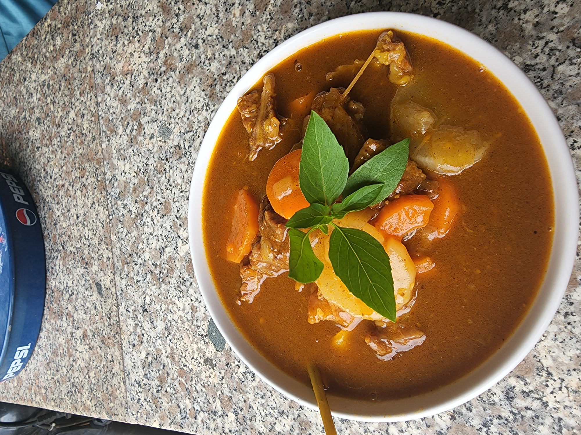 A white bowl of beef stew with carrots and potatoes sits on a speckled granite countertop. The stew has a rich, brown broth and is garnished with fresh green basil leaves. A light-colored wooden skewer is visible in the bowl, resting against the side. In the background, a blue and silver Pepsi tray is partially visible, its logo clearly displayed. The scene is likely set in a casual dining establishment or outdoor eatery, possibly a roadside restaurant. The question of the specific location in Phan Thiet Ward, Vietnam is not answerable based on the provided image alone, as there are no identifying landmarks or contextual clues that pinpoint a precise neighborhood or establishment.