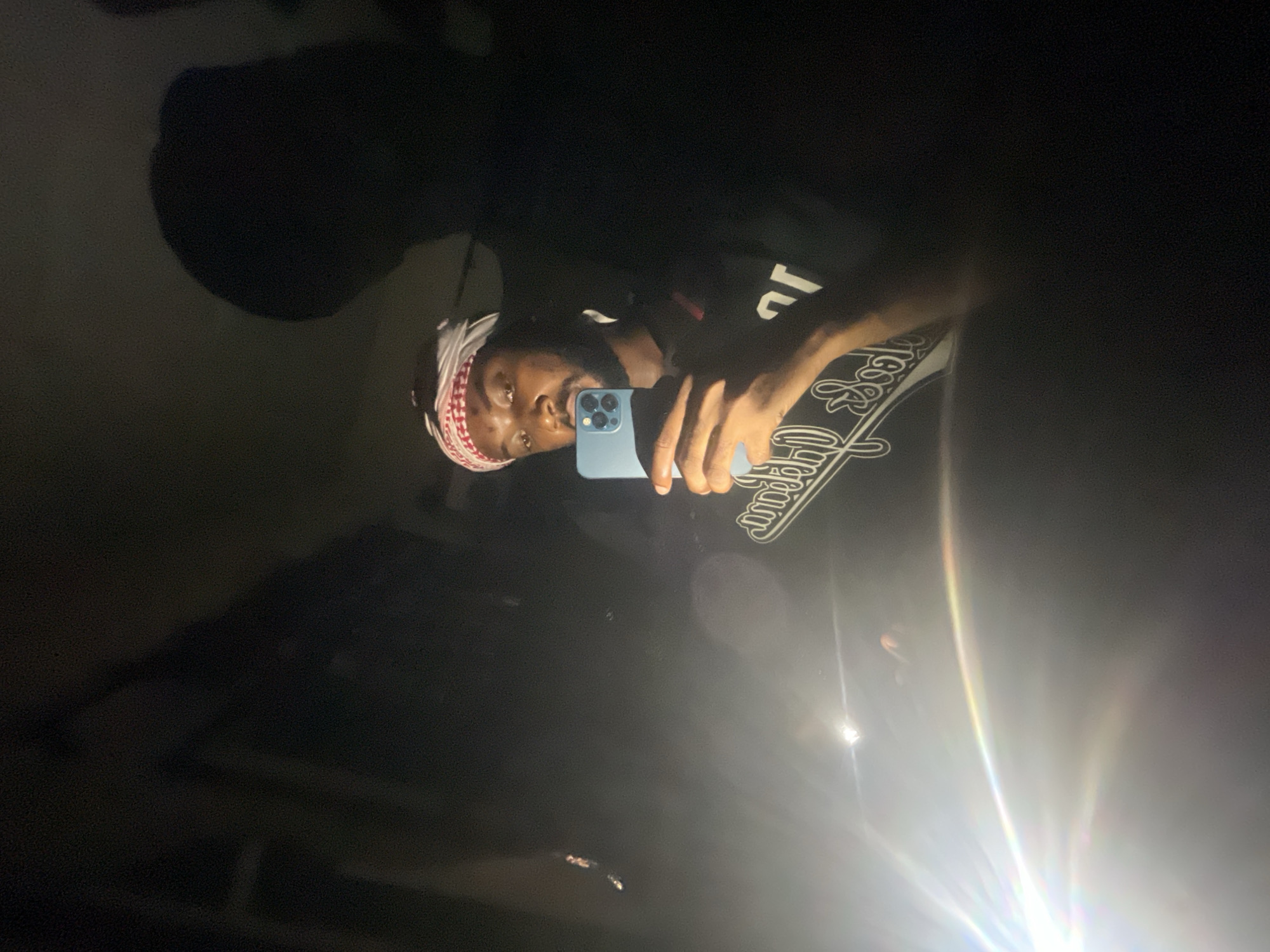 A dark-skinned person wearing a head covering is holding a blue smartphone, presumably taking a selfie. The person is wearing a black shirt with white writing on it. The background is very dark with some light flare. The location is unknown and there is no information to determine that the location is Lokoja, Nigeria.