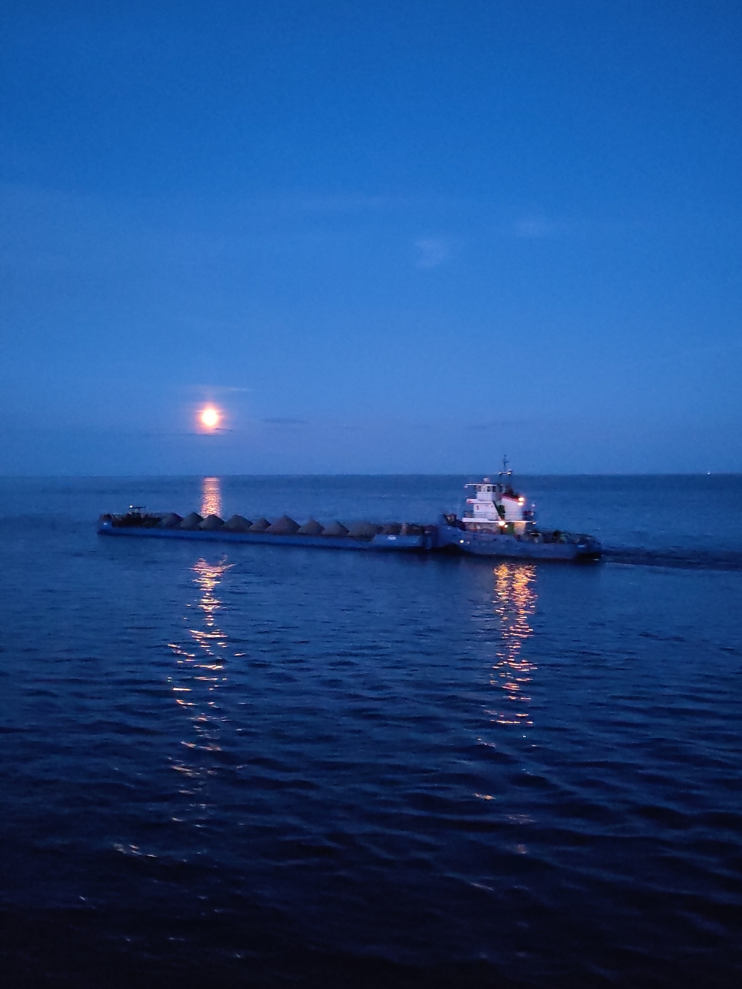 A low-angle, long-shot captures a barge being towed by a tugboat on a body of water under a twilight sky. The barge is laden with what appears to be aggregate material. The tugboat is situated at the rear, with visible lights and a superstructure.
Above the horizon, a luminous orb, presumably the moon, is faintly visible, reflecting on the water's surface, creating a vertical path of light. The water is moderately choppy, with small waves generating visible reflections. The sky transitions from a dark blue at the top to a lighter hue near the horizon. The overall color palette is cool, dominated by blues and grays, with the highlights from the lights creating warm accents. The location is Unknown, Unknown.