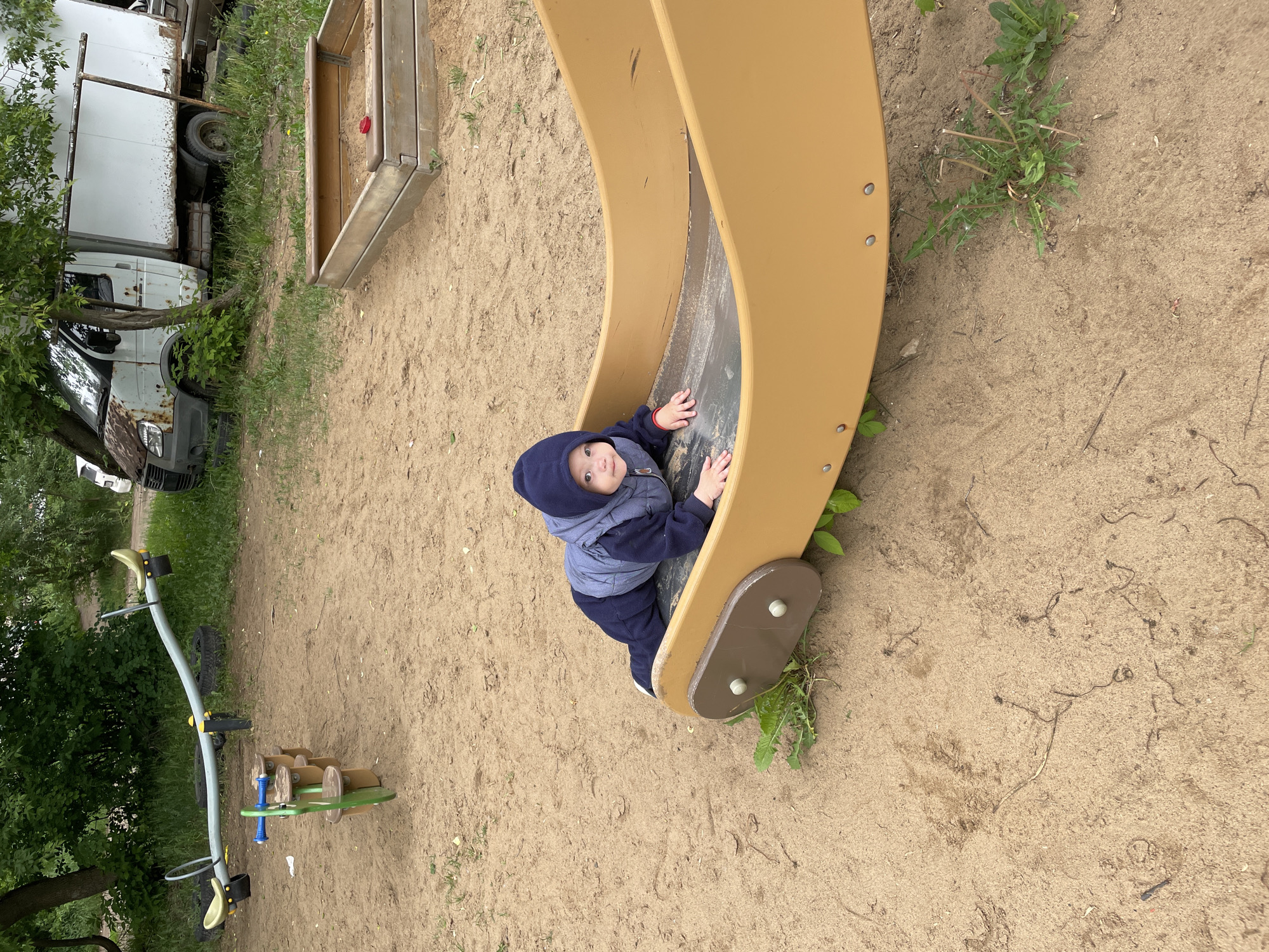 This image captures an outdoor playground scene in Perm, Russia, under an overcast sky. In the foreground, a young child, appearing to be an infant or toddler, is positioned on its stomach on the lower portion of a mustard-yellow and dark-grey playground slide, looking directly towards the viewer. The child is wearing a dark blue hooded garment over a grey vest and dark blue trousers.
The playground features extend into the midground:
* A light blue seesaw with yellow seats and black handgrips is visible to the left of center.
* A green and brown spring rider stands in front of the seesaw.
* To the upper right, a wooden sandbox frame is partially visible, containing sand and a small red object.
The ground across the entire visible area consists primarily of light brown sand, with interspersed patches of green grass and weeds, particularly around the base of the playground equipment.
In the background, behind the playground structures and a line of dense green foliage, two dilapidated white box trucks are parked. The truck on the left shows significant rust on its cab and body panels, characteristic of a GAZelle model. The truck on the right is also white with visible rust and appears to be a similar utility vehicle. The presence of these aged vehicles suggests an adjacent industrial or neglected area.