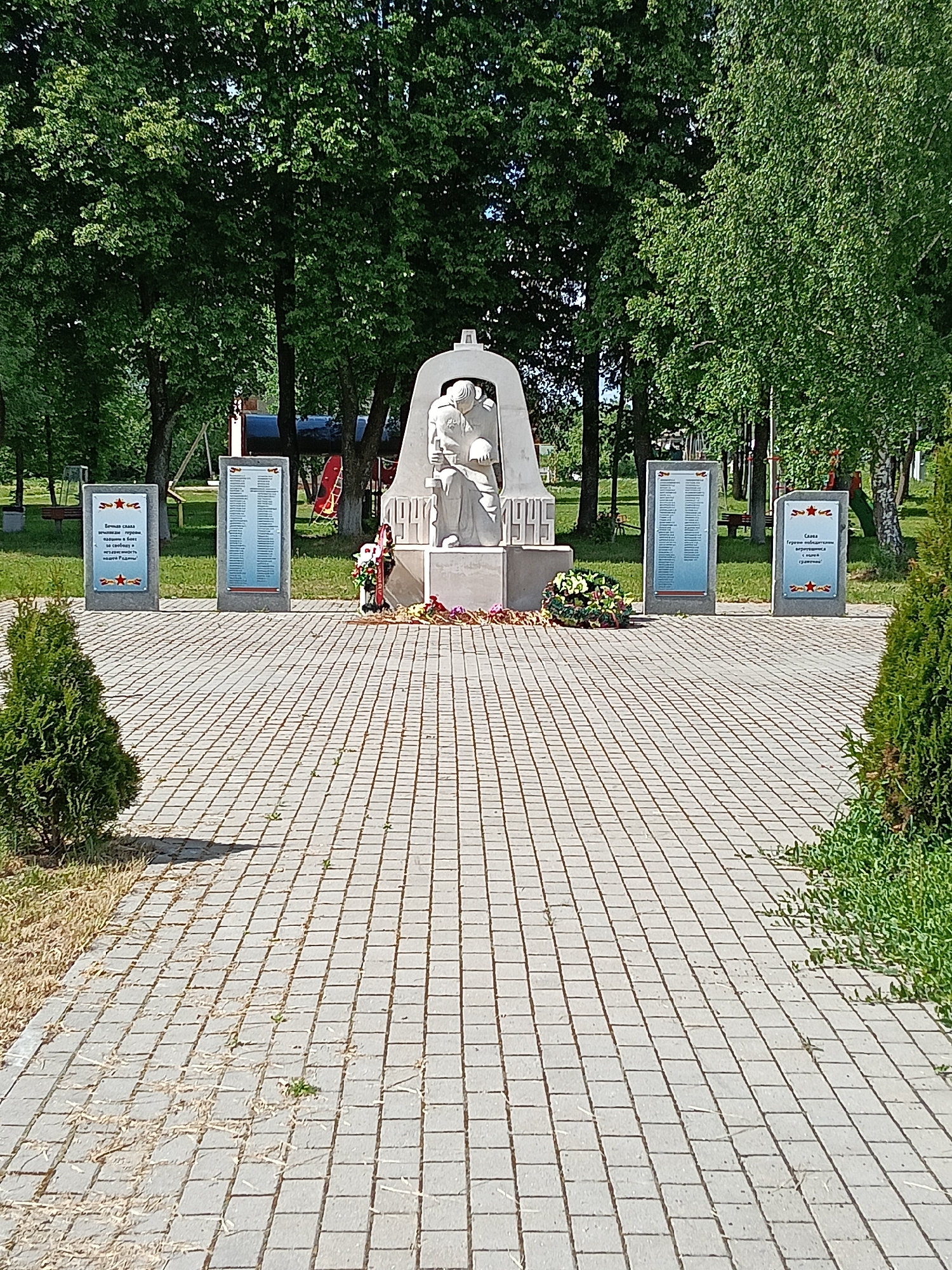 The image captures a memorial complex in the city of Красный Маяк, Russia. The focal point is a white marble sculpture depicting a kneeling figure, possibly a soldier or a mourner, head bowed and holding a helmet. The sculpture is set within a concrete archway, with the number "1945" inscribed on its base.
Surrounding the central sculpture are four plaques on stands, presumably listing names or commemorating events. Flowers and wreaths are laid at the base of the sculpture, indicating the space is a place of remembrance.
The memorial is situated on a paved area, with small trees and greenery providing a backdrop. The sky is not visible, but the lighting suggests it is daytime. In the background, playground equipment can be seen suggesting the memorial is located in a park.