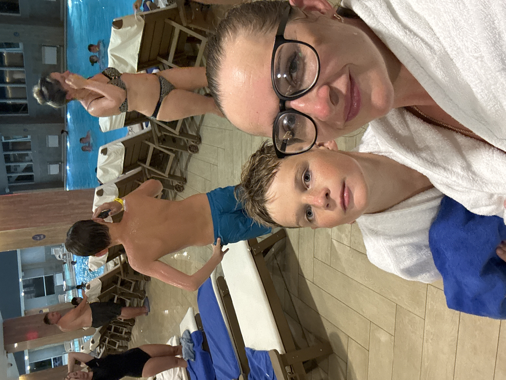 An indoor swimming pool area in Anapa, Russia, features a woman in a black swimsuit and a man in black shorts standing near lounge chairs, while a woman in a bikini walks past another lounge chair. In the foreground, a boy in blue shorts faces the camera, and a woman wearing glasses and a white robe takes a selfie with the boy. The pool water is blue, and lounge chairs are arranged around the pool. The floor is tiled, and the walls are decorated with columns.