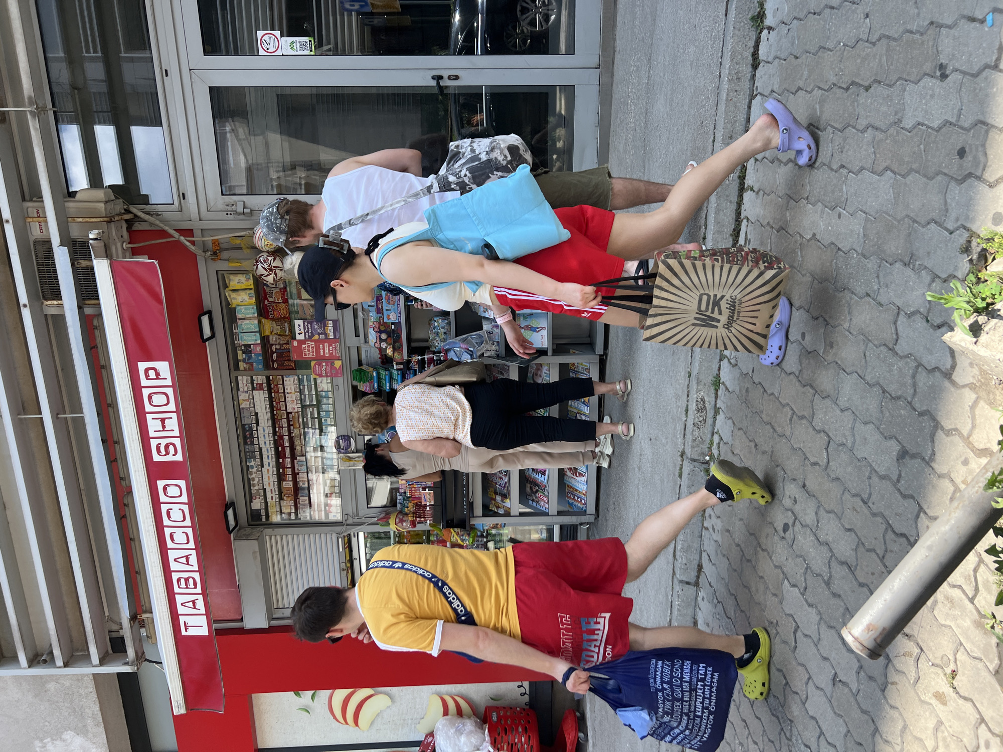 In Budva, Montenegro, four individuals walk past a tobacco shop. The shop's red awning displays "TABACCO SHOP" in white lettering. A display window showcases various products. The first person, a man in a yellow t-shirt and red shorts, carries a blue shopping bag. He wears lime green Crocs. Behind him, a woman in a white tank top and red shorts, with a black baseball cap, carries a light blue tote bag and a brown paper bag. She wears light purple Crocs. To her right, a man in a white tank top and olive green shorts carries a marble-patterned bag. Further to the right, a woman in a beige top and black pants carries a tan purse. The sidewalk is paved with gray interlocking stones, and a metal pipe lies on the ground in the foreground.