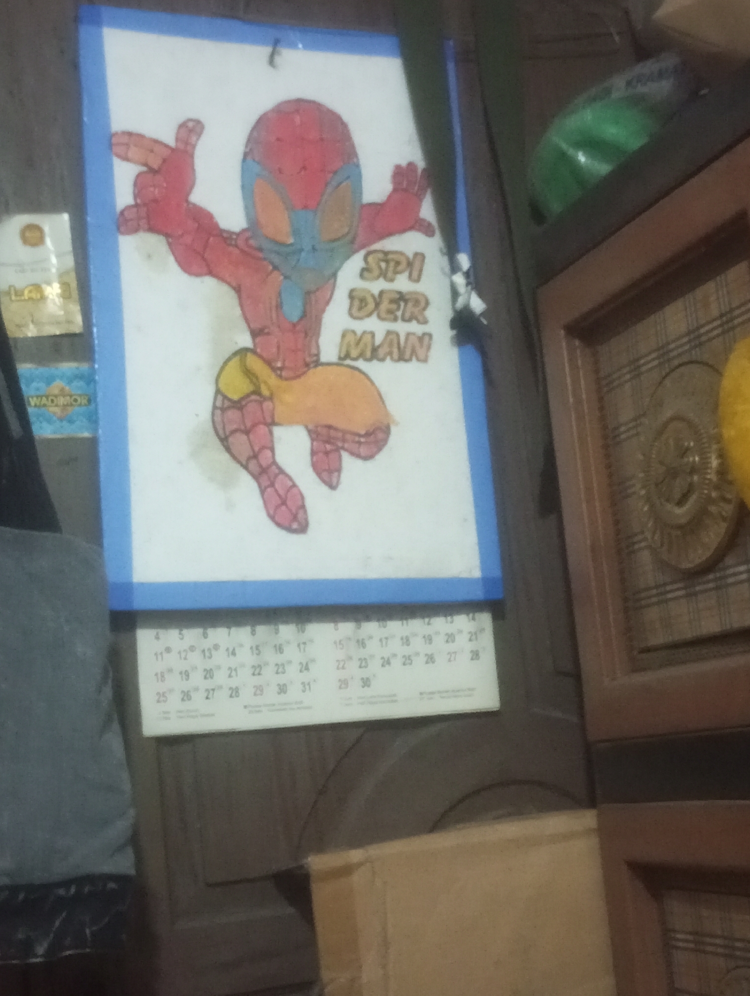 The image depicts a close-up of a Spiderman drawing on a whiteboard, with a calendar visible below it. The drawing shows Spiderman in a dynamic pose, with his red and blue costume. The text "SPIDER MAN" is written next to the drawing. The calendar appears to be for the month of March. In the background, there is a brown cabinet with a decorative circular emblem. To the left of the whiteboard, there are two small labels, one with "LAPI" and another with "WADIMOR". The overall scene suggests an indoor setting, likely a home or an office. The image is blurry and poorly lit, making it difficult to discern finer details. It is not possible to determine the precise location within Cilegon, Indonesia from the visual information provided.
