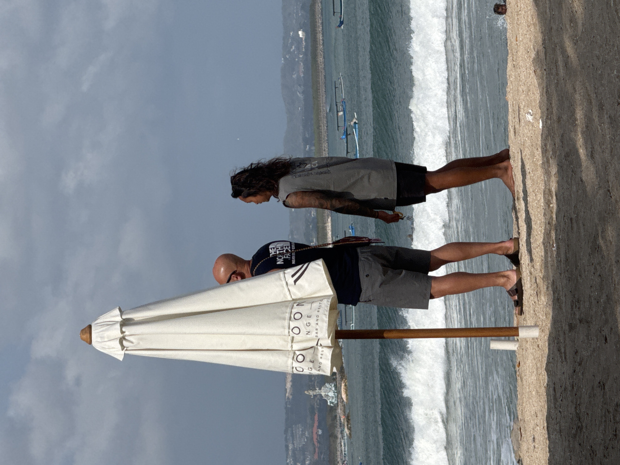 Two people stand on a sandy beach in Kuta, Indonesia, near the ocean with waves. In the foreground, a tall, closed white umbrella with a brown wooden pole stands partially obscuring a bald man wearing sunglasses, a dark t-shirt with "The North Face" logo on the back, and grey shorts. He is looking out at the ocean. To his right, a person with long, dark, wavy hair, wearing a grey sleeveless t-shirt and black shorts, also looks out at the ocean. Their arms are bare, and the person on the right has tattoos on their left arm. In the background, several narrow boats with canopies are visible on the water. Further in the distance, a distinctive white monument with a statue is visible on a rocky outcrop in the ocean. The sky is overcast with scattered clouds. The sand in the foreground is textured, with some shadows cast by unseen objects.