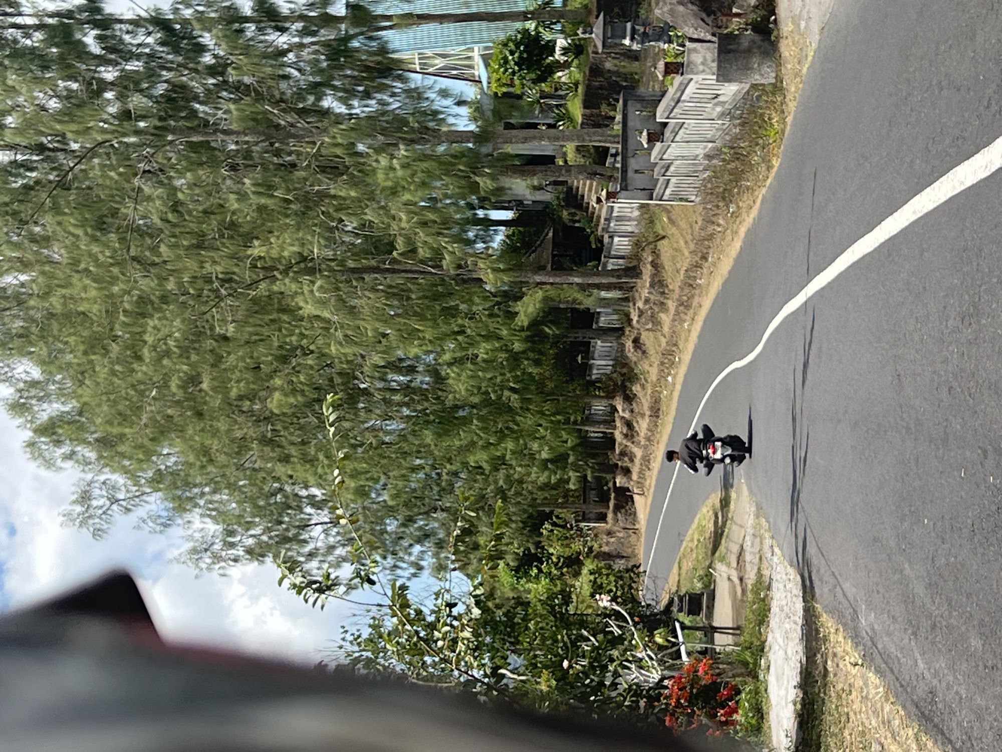 The image shows a paved road winding through a rural area in Kintamani, Indonesia. A person is riding a motorcycle away from the camera on the right side of the road. The road is lined with trees and a stone wall. The sky is partly cloudy. There are buildings visible in the background.