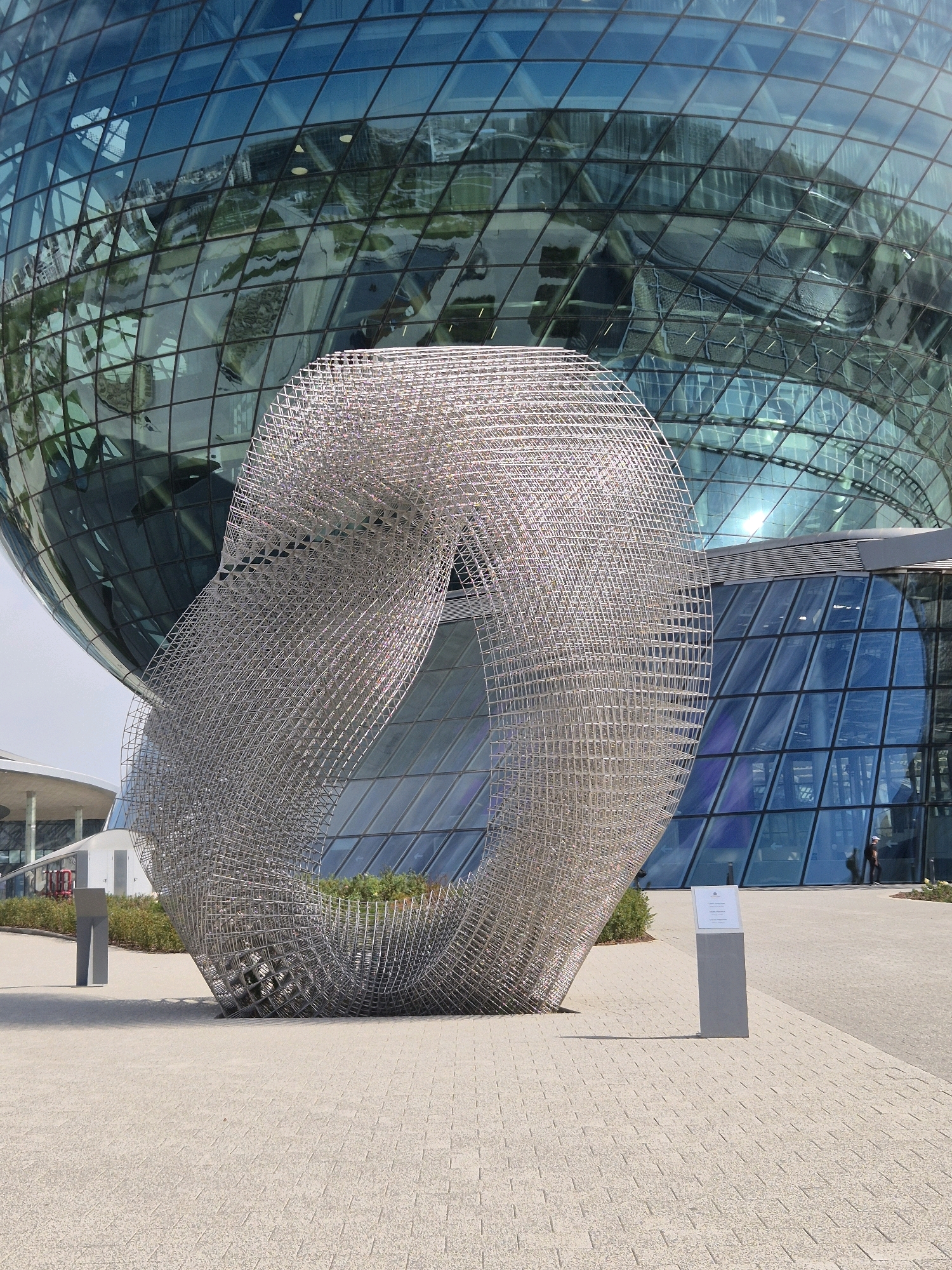 The image depicts a large, metallic, toroidal sculpture in front of a spherical glass building. The sculpture is composed of a grid-like structure, creating a hollow interior with a woven appearance. The glass building, which reflects the sky and surrounding environment, dominates the background. The scene is set outdoors on a paved plaza, with some greenery visible behind the sculpture and to the left of the building. Two rectangular grey posts are present on the plaza, one to the left and another to the right of the sculpture, with a sign on the right post. A single person is visible in the background, walking near the entrance of the glass building. The location is identified as Astana, Kazakhstan.