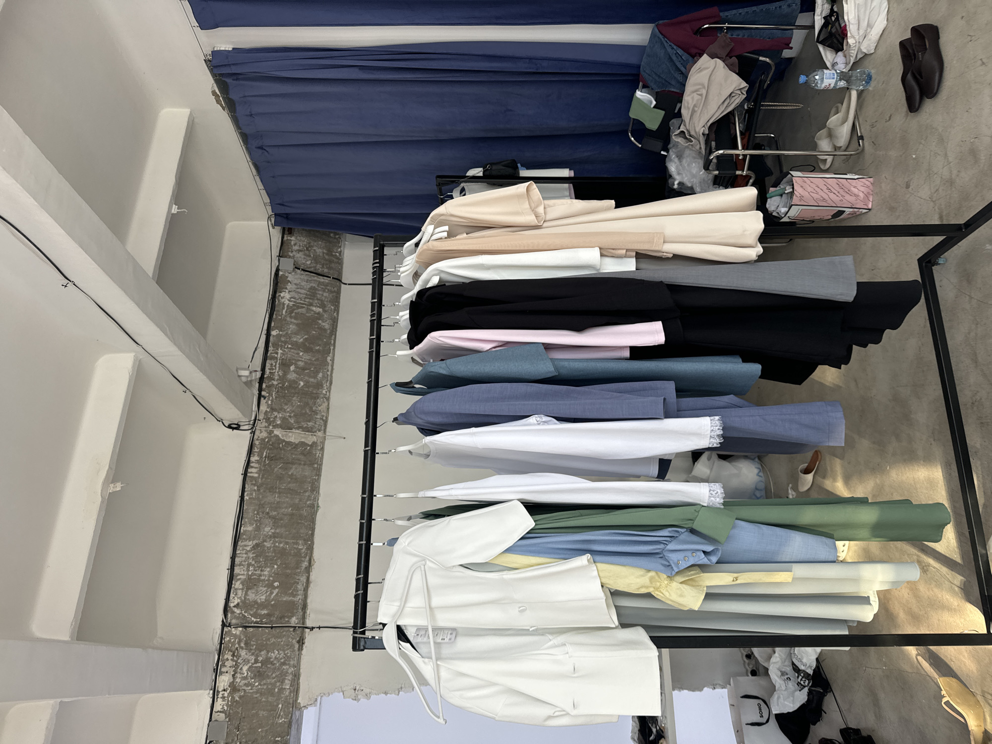 The image depicts a clothing rack indoors, likely in a retail or studio setting, possibly Krasnoyarsk, Russia. The rack holds an assortment of garments in various colors, including beige, white, black, pink, blue, green, and yellow. The clothes appear to be primarily shirts, blouses, and potentially jackets. A blue curtain hangs in the background. Adjacent to the rack, assorted items are visible, including a shopping cart with clothes, a water bottle, and shoes. The ceiling is white and appears to have structural support beams.
