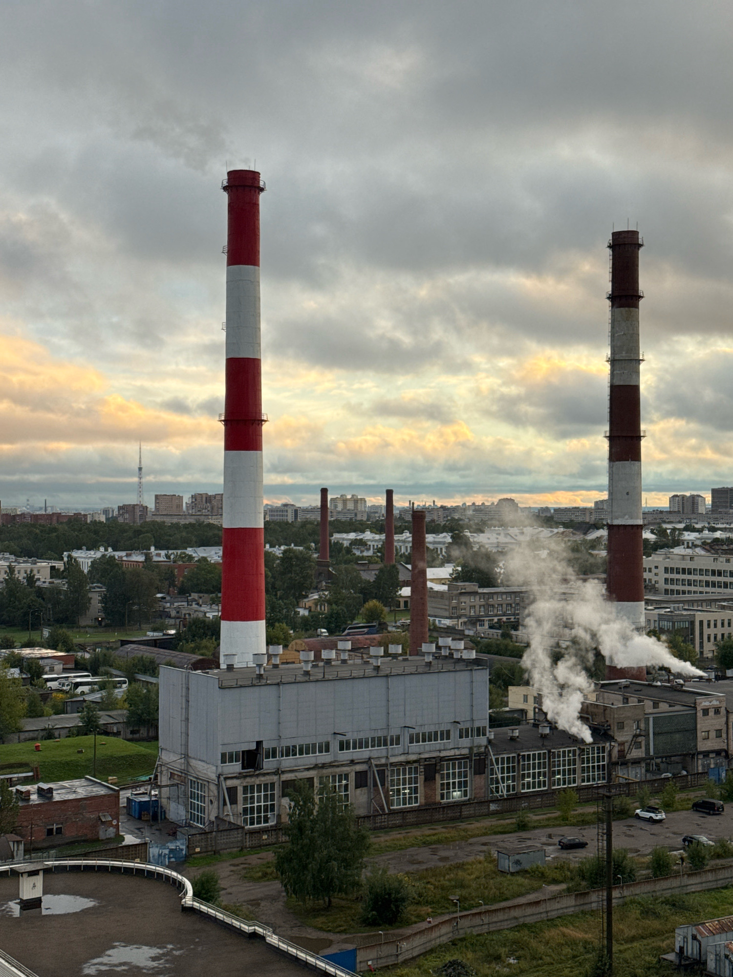 The image shows an industrial area with two large smokestacks emitting smoke against a cloudy sky. The smokestacks are tall and cylindrical, with red and white bands on one and red and grey bands on the other. In the foreground, there is a large industrial building with a metal roof and numerous windows. Surrounding the building are smaller structures, roads, and patches of green vegetation. In the background, a city skyline with apartment buildings and a distant tower is visible. The overall scene suggests an active industrial site within a city.