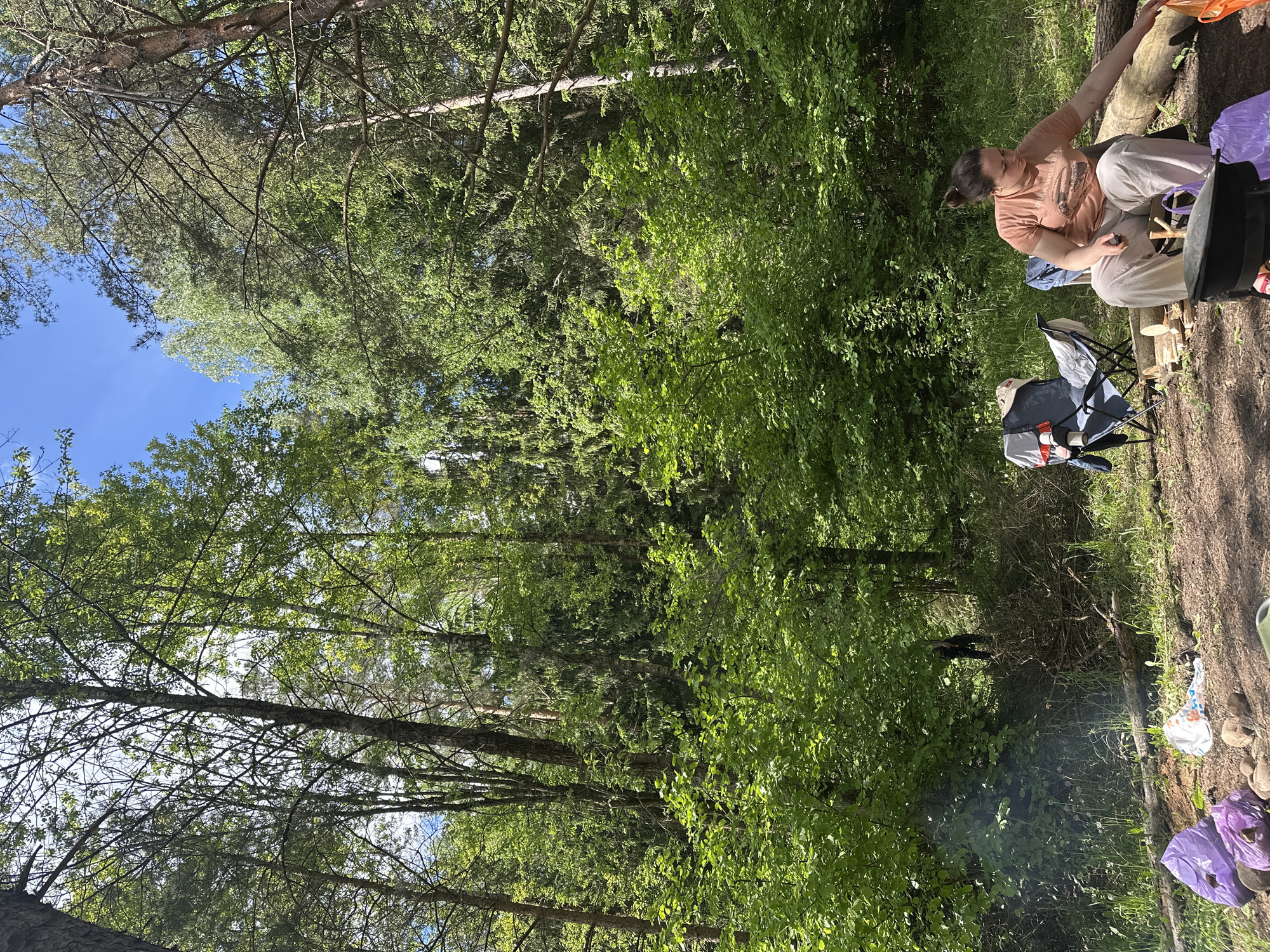 The image shows a campsite in a forested area in Юзуфоўскі сельскі Савет, Belarus. A woman is seated near a fallen log, with a folding chair and other camping gear nearby. Trees dominate the background, and the sky is visible through the canopy. There is some smoke arising near the bottom of the image. There is evidence of human presence, including trash bags and various other items associated with camping.
