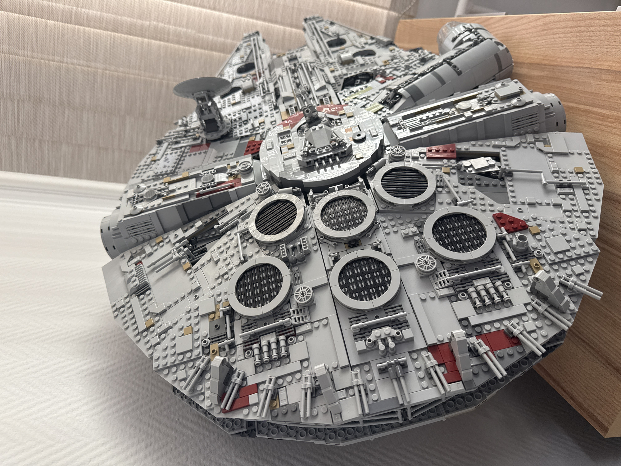 The image depicts a detailed LEGO model of the Millennium Falcon. The model is predominantly gray and intricately constructed with various LEGO bricks. It features recognizable elements such as the cockpit, radar dish, and engine vents. The model is placed on a surface; on the left is a white surface and on the right a wooden surface. The background consists of a beige wall. The location is not apparent from the image.