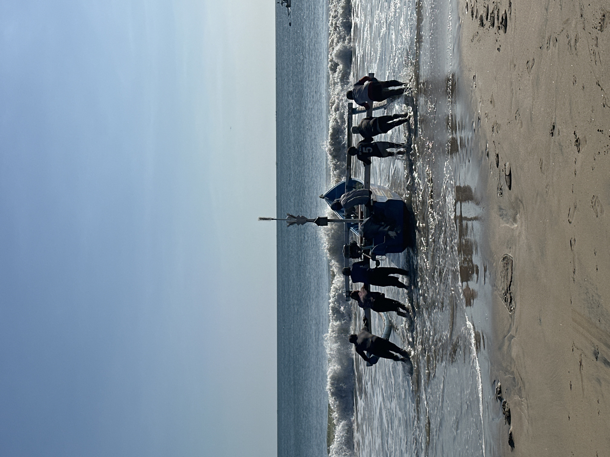 A group of seven men are working together to pull a blue and white fishing boat out of the ocean and onto the sandy shore. The men are positioned on either side of the boat and are pushing and pulling it with their hands and bodies. The boat is partially submerged in the water, with waves crashing around it. The sky is clear and blue, and the sun is shining brightly. The beach is made of light brown sand, and there are some footprints visible in the sand near the water's edge. In the distance, the ocean stretches out to the horizon, with a few other boats visible. This scene likely depicts fishermen in Kuta, Indonesia, engaged in the daily ritual of bringing their boats ashore.