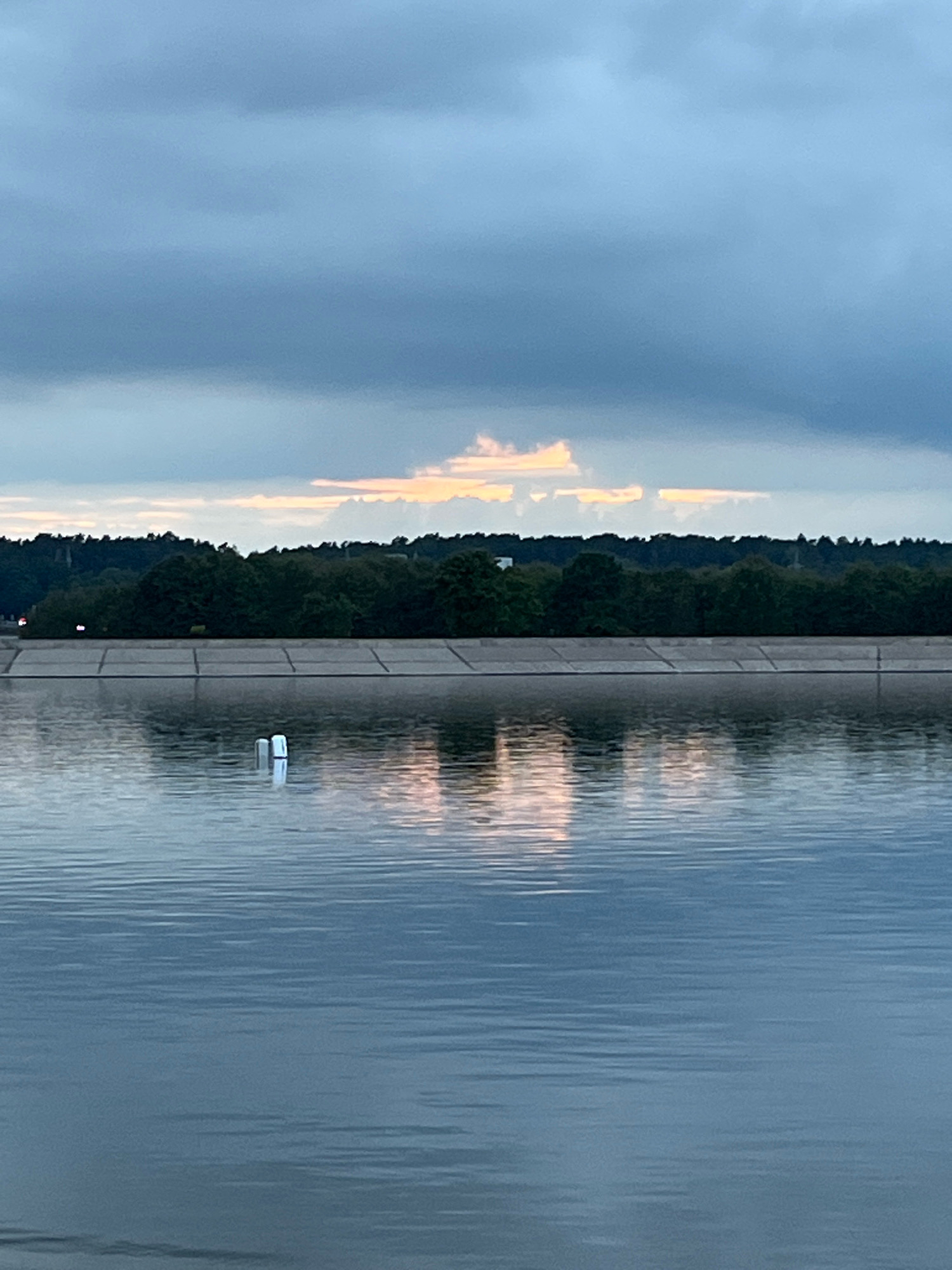 The image depicts a body of water, possibly a reservoir or a large pond, in Kaunas, Lithuania. The water's surface is calm with gentle ripples reflecting the sky and the opposite bank. In the foreground, two white cylindrical buoys are partially submerged in the water, with their reflections also visible.
The middle ground features a concrete embankment or wall, extending horizontally across the frame. This structure appears to be made of large, rectangular blocks. Beyond the embankment, a dense line of trees forms the treeline of the opposite bank, creating a dark green mass against the sky.
The sky is overcast with dramatic clouds, predominantly in shades of gray and blue. Near the horizon, a break in the clouds allows a sliver of warm, soft orange and pink light to penetrate, suggesting either sunrise or sunset. This light is reflected on the water's surface, adding a touch of color to the otherwise muted tones of the scene. The overall impression is one of a tranquil, perhaps early morning or late evening, landscape.