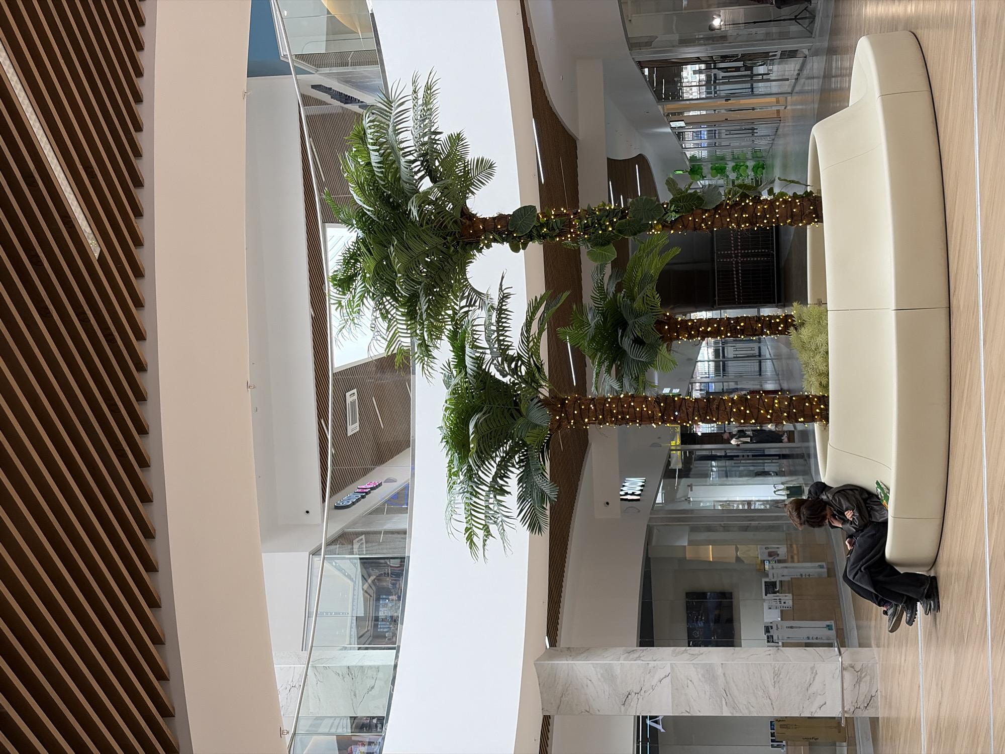 The image displays the interior of a brightly lit, modern shopping mall or large public atrium in Izhevsk, Russia. Two individuals are seated on a long, curved, light beige bench positioned on the right side of the frame. One person, with dark hair and dark clothing, is leaning forward, seemingly engrossed in something on their lap or a personal device. The other person is partially obscured behind them.
Central to the scene are two tall artificial palm trees with vibrant green fronds. Their trunks are elaborately wrapped with strands of small, glowing string lights, adding a decorative and possibly festive touch to the environment.
The architectural design is contemporary, characterized by clean lines and a mix of materials. On the far left, a large slatted wooden panel runs diagonally, creating a striking texture. Smooth, light-colored walls and pillars contribute to the open and airy feel. Transparent glass railings and panels extend through the space, reflecting other parts of the interior, including distant sections of the mall with shop windows. The floor beneath the bench is light-colored and wood-patterned, while areas in the foreground feature marble-patterned surfaces.
In the background, visible through the glass barriers, a vertical black sign with white text clearly displays "300M". The scene depicts a calm moment of rest within a spacious and aesthetically designed public indoor setting.