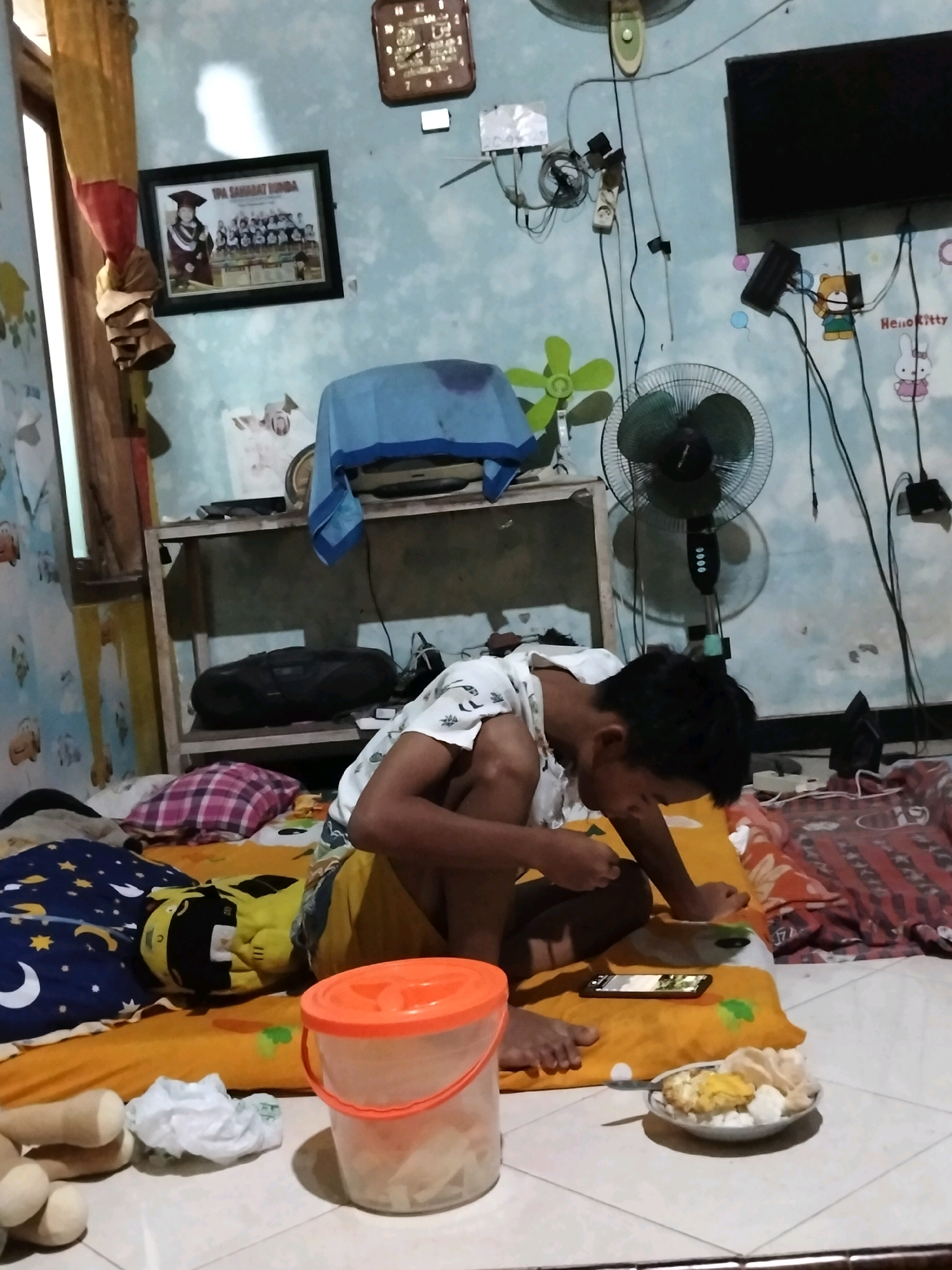 The image depicts an interior scene, likely a bedroom, with a young male sitting on the floor. He is wearing a white t-shirt and appears to be eating. In front of him is a plate of food, a mobile phone, and a plastic container with an orange lid. The room is sparsely furnished with a bed, a shelf with a stereo system, and various objects on the walls, including a framed picture and an electronic fan. The walls are painted a light blue color and adorned with decorative stickers, including Hello Kitty characters. The overall impression is one of a modest, lived-in space. The prompt mentions the location as Puskesmas Keboan, Indonesia, but this information cannot be visually confirmed from the image itself.