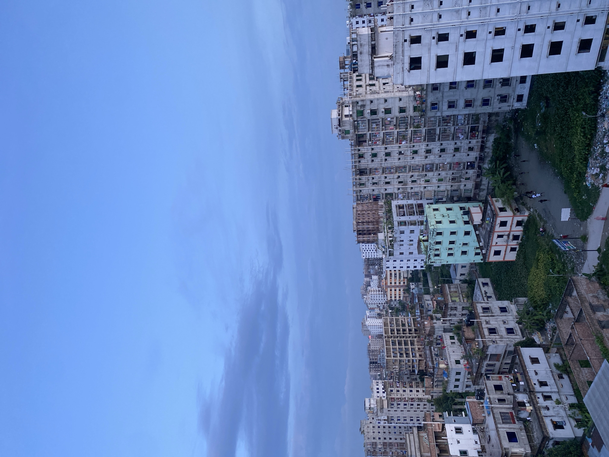 This is an aerial view of a densely populated urban area in Dhaka, Bangladesh, characterized by numerous multi-story buildings, many appearing to be under construction or in various states of repair. The sky is overcast with a soft blue hue, suggesting it might be early morning or late evening. The buildings are a mix of concrete structures, some with exposed rebar, indicating ongoing construction. Others are complete with balconies and windows, some of which have clothing hanging out to dry. There are patches of green vegetation interspersed between the buildings, and some areas appear to be filled with debris or refuse. A few people are visible in the lower parts of the image, walking on unpaved paths or open ground. One area shows a group of individuals gathered, possibly playing or socializing. The overall impression is one of a rapidly developing urban landscape with a high population density.
