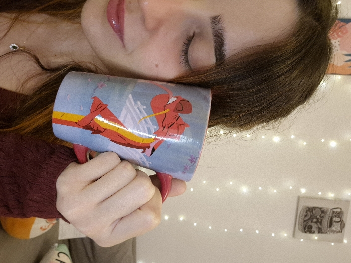 A person with closed eyes, glossy lips, and long brown hair cradles a light blue mug with a red dragon design. They are wearing a maroon-colored sweater. The background includes a string of warm-toned fairy lights and a small, framed black and white picture. The overall composition is rotated and tilted, presenting a close-up, personal perspective.