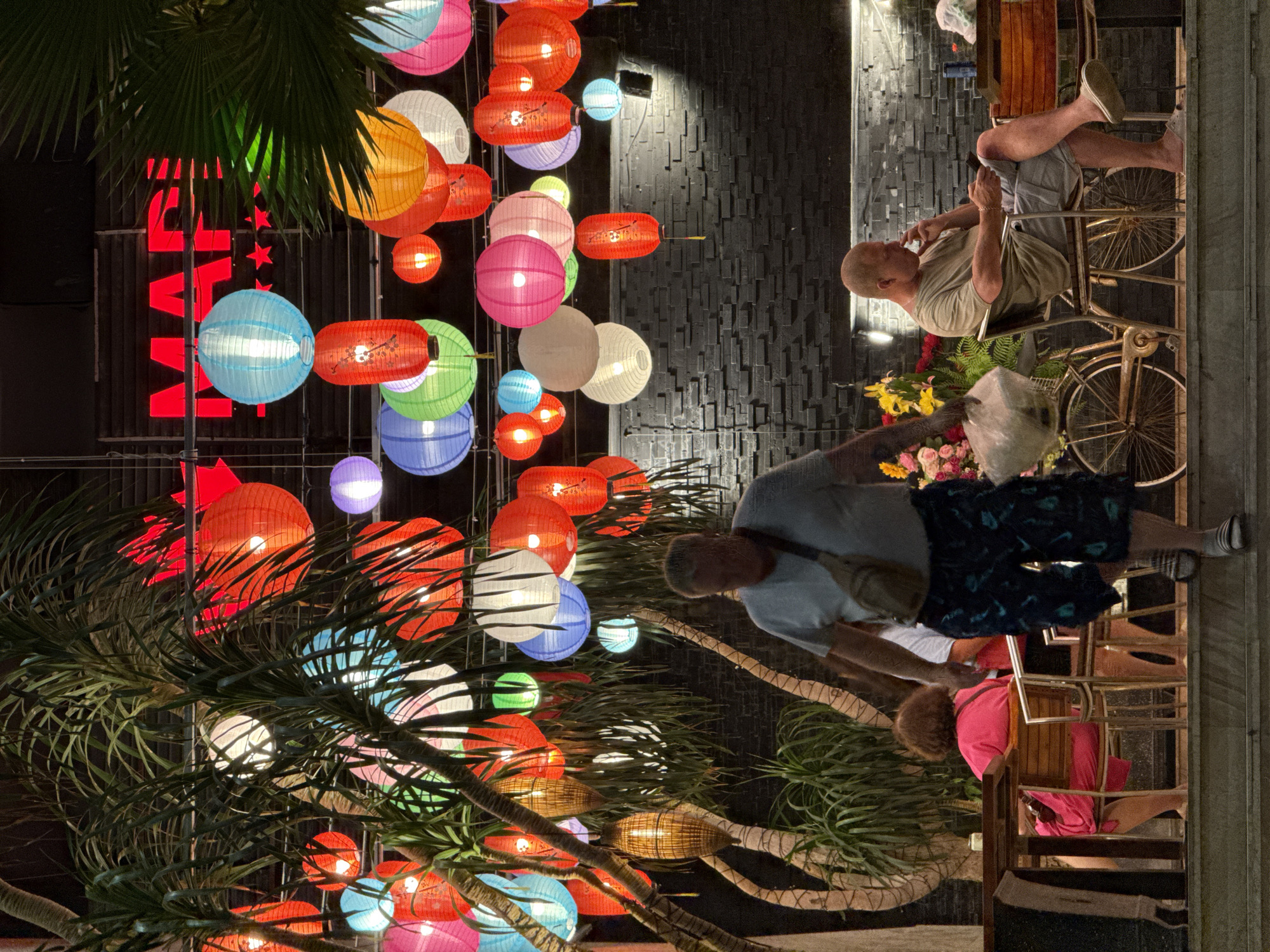 The image captures a vibrant nighttime street scene in Nha Trang, Vietnam, illuminated by numerous colorful lanterns and neon signs. The perspective is tilted, making the scene appear rotated. Overhead, a dense canopy of palm fronds intertwines with dozens of glowing paper lanterns of various shapes and sizes. These lanterns display a rich palette of red, blue, green, pink, orange, and white, some featuring subtle patterns.
In the midground, a large red neon sign spelling "MAP" with accompanying stars is visible against a dark, textured wall. Below the sign and lanterns, several individuals are present. One man with a shaved head sits relaxed on a traditional Vietnamese cyclo or bicycle taxi, engrossed in his phone. He wears a light-colored t-shirt and shorts, with a gold-colored bracelet visible on his wrist.
To the right, another man, also with light skin, stands with his back partially turned, holding a white plastic bag. He wears a light t-shirt and dark, patterned shorts. Nearby, a vibrant display of yellow, pink, and red flowers in a pot adds another splash of color to the scene. Further in the background, a person with reddish-brown hair is seated at a table, wearing a bright pink top. The dark, patterned wall provides a contrasting backdrop to the lively scene. The overall ambiance suggests a bustling evening street or restaurant area, characteristic of a lively tourist or local district.