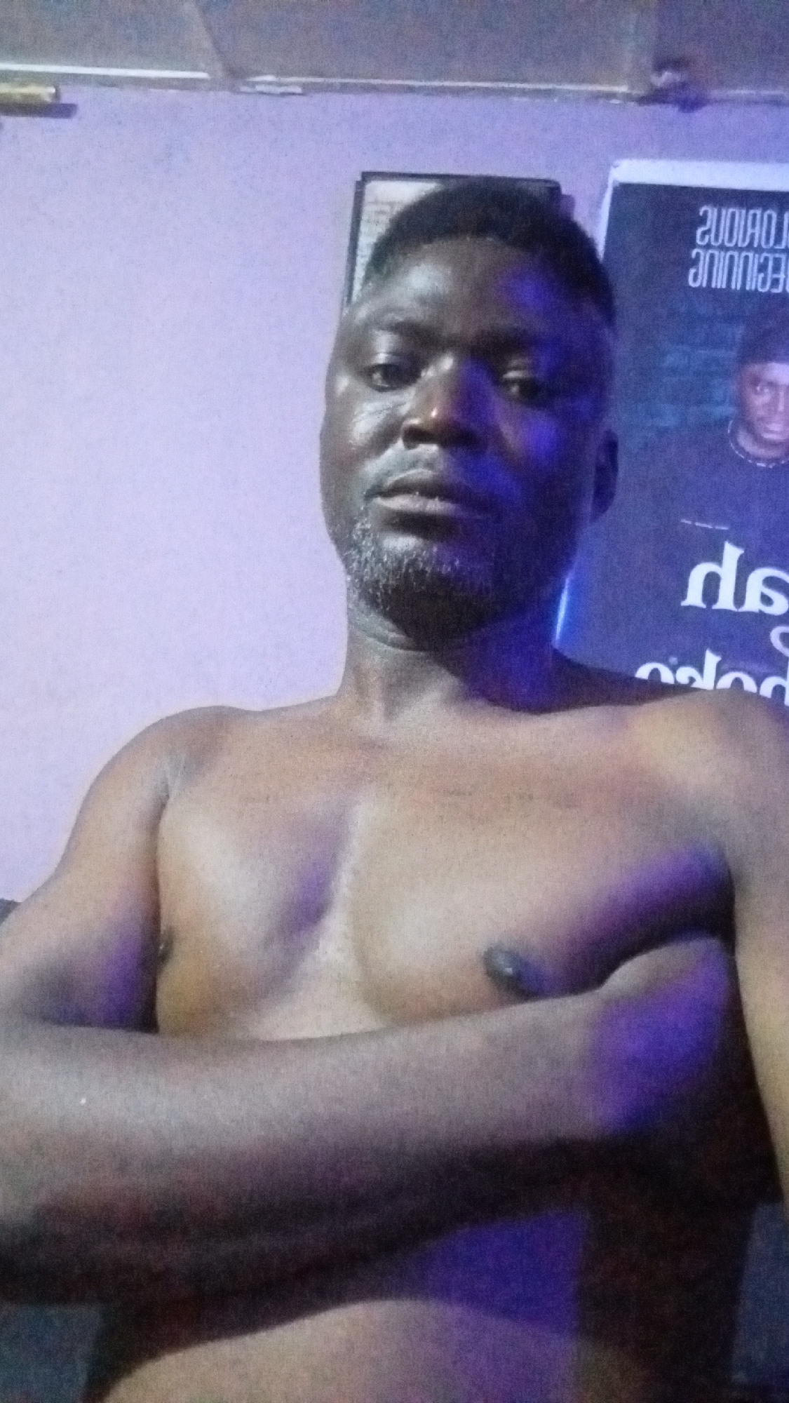A shirtless man with short, dark hair and a graying beard is the central subject of the image. He is looking directly at the camera with a neutral expression, his arms crossed over his chest. The man's skin tone is dark, and his chest muscles are visible.
He is positioned indoors against a light-colored wall, which appears to be a shade of purple or lavender, possibly due to ambient lighting. Behind him, to the viewer's left, a dark framed object, possibly a picture or mirror, is visible on the wall. To the viewer's right, a large poster or banner hangs on the wall.
Visible text on the poster is mirrored, making it appear backward. Partially decipherable words include "SOUЯO⅃O" (likely "GLORIOUS" mirrored) and "ƎUИIИИIƎ". Below these, smaller mirrored text like "qs" and "pələc" can be seen. A small image of another person's head is also visible on the poster. The overall lighting has a cool, blue-purple hue, suggesting artificial light in the room. The setting appears to be a casual indoor space.