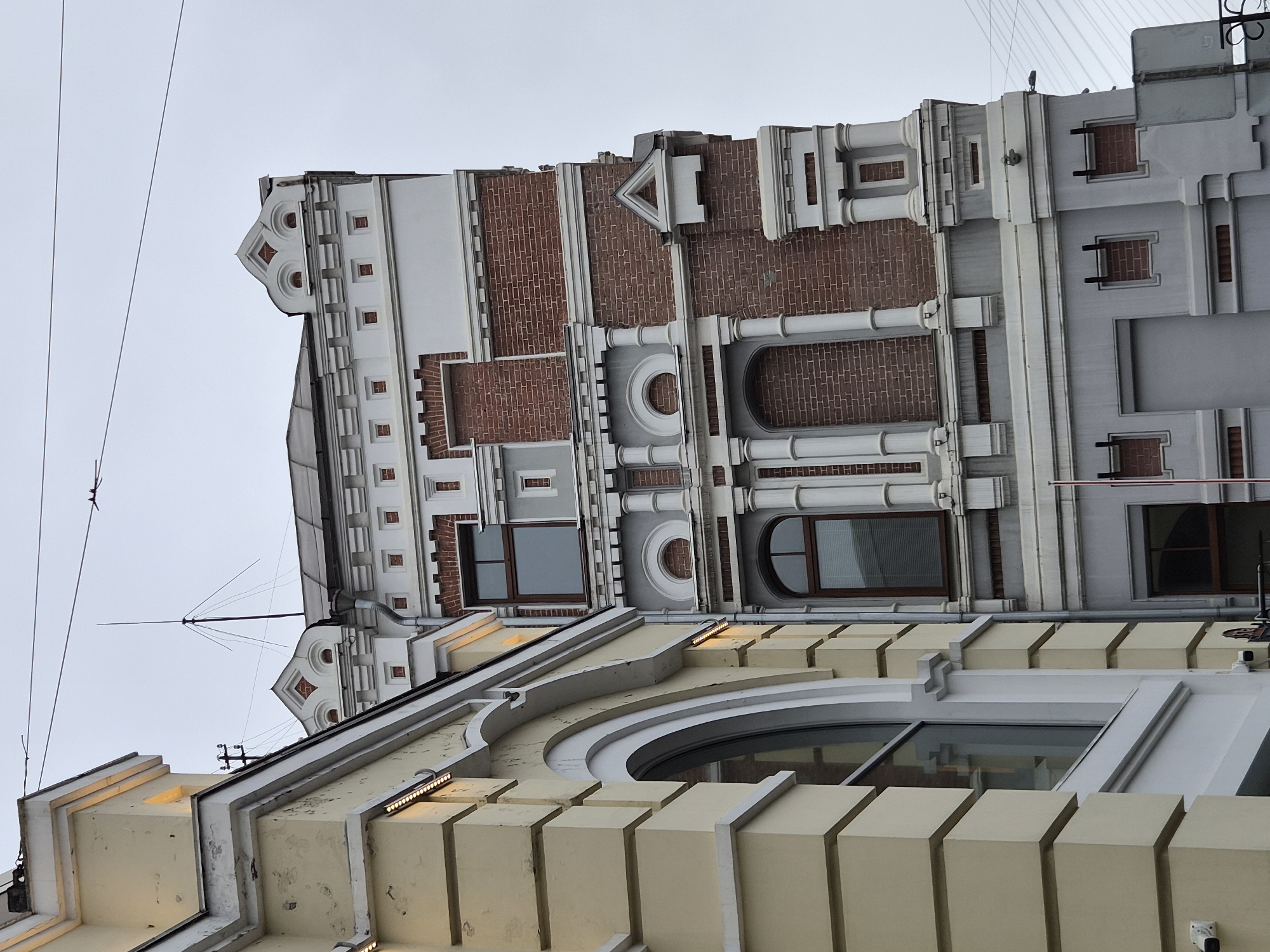 The image shows an exterior view of two adjacent buildings in Vladivostok, Russia, against a cloudy sky. The building on the left is lighter in color, possibly yellow or cream, with a curved archway and horizontal banding details. It features a large arched window and an external security camera mounted near the bottom left corner.
The building on the right is taller and more complex in design, featuring a contrasting facade of red brick and white architectural elements. This building has a more ornate appearance with multiple levels, decorative cornices, recessed brick sections, and columns. Windows are visible on various levels, some with dark frames and blinds. A tall antenna or lightning rod extends from the rooftop of this building. Electrical wires are strung across the sky above the buildings. The overall scene depicts an urban architectural setting.